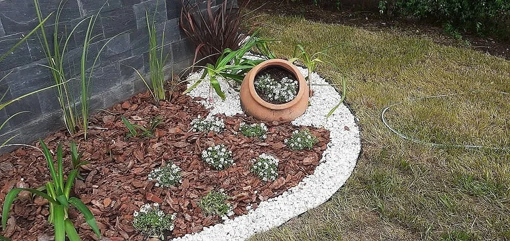 Jardín decorativo con plantas, flores blancas y una olla de barro con agua.