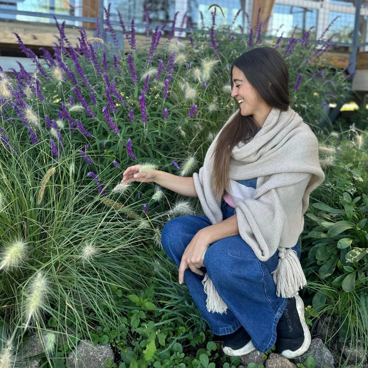Mujer sonriendo mientras tocas plantas en un jardín con flores moradas y verdes.