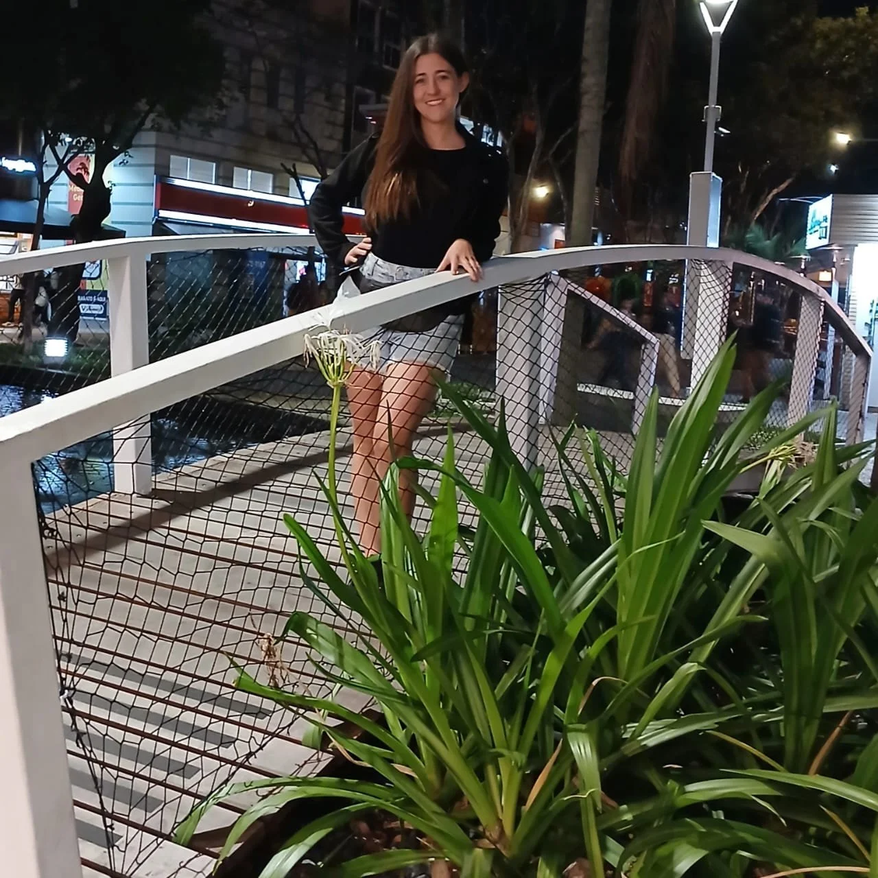 Una mujer sonriente posa sobre un puente peatonal en una ciudad de noche, con árboles, luces y edificios iluminados alrededor.