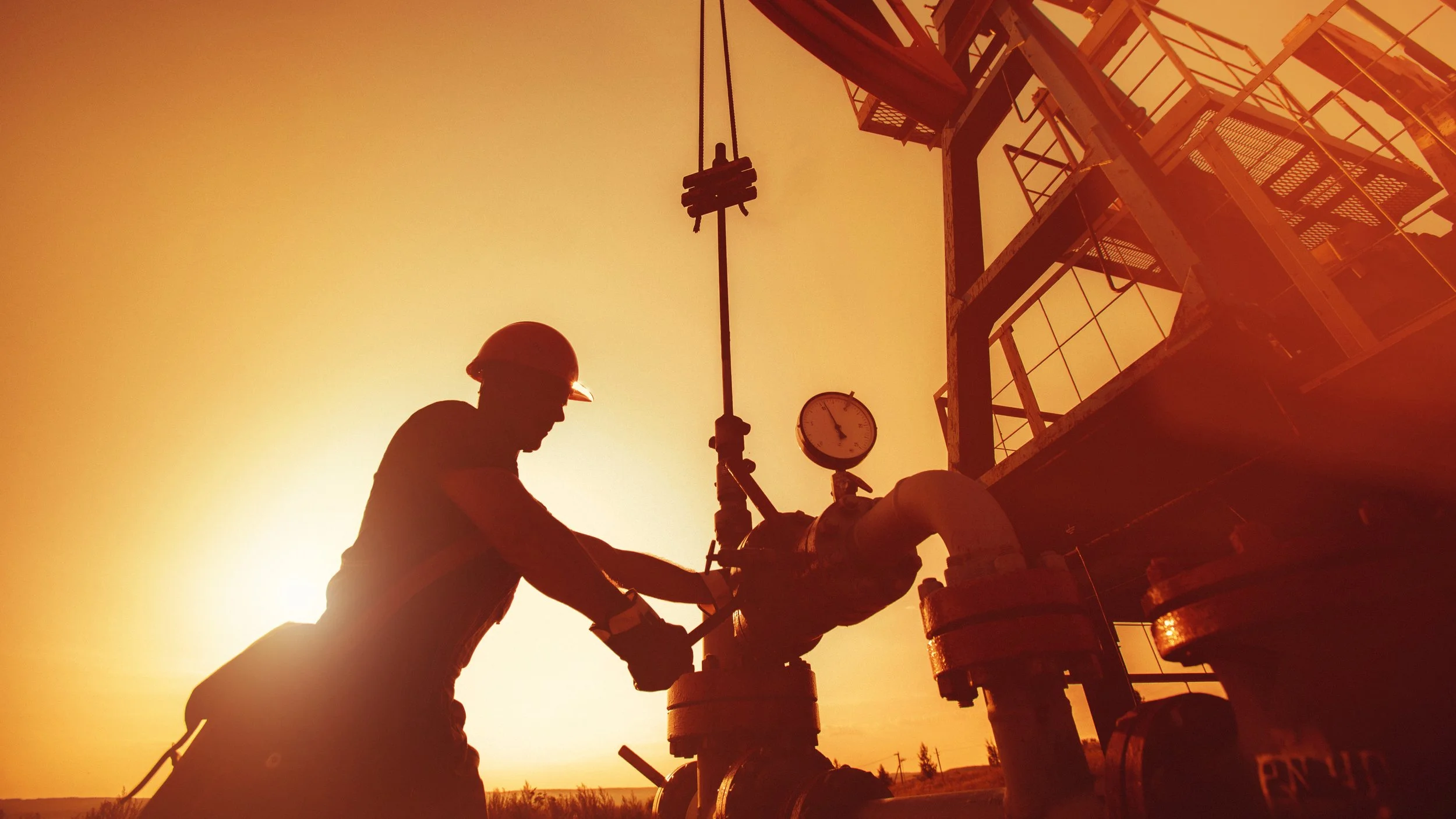 Silhouette of an oil or gas worker wearing a hard hat, working on industrial piping during sunset or sunrise, with a large industrial structure and a round pressure gauge visible in the background.