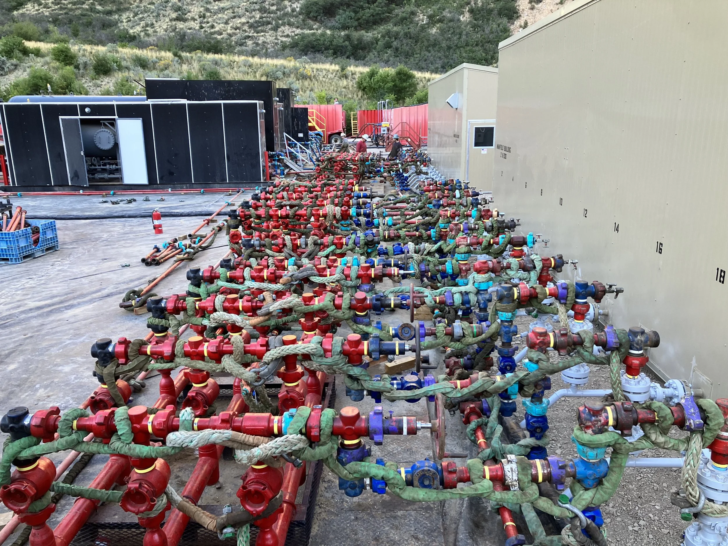 Pipes, hoses, and valves are laid out on the ground at a construction site, with a few construction workers working in the background and hills in the distance.