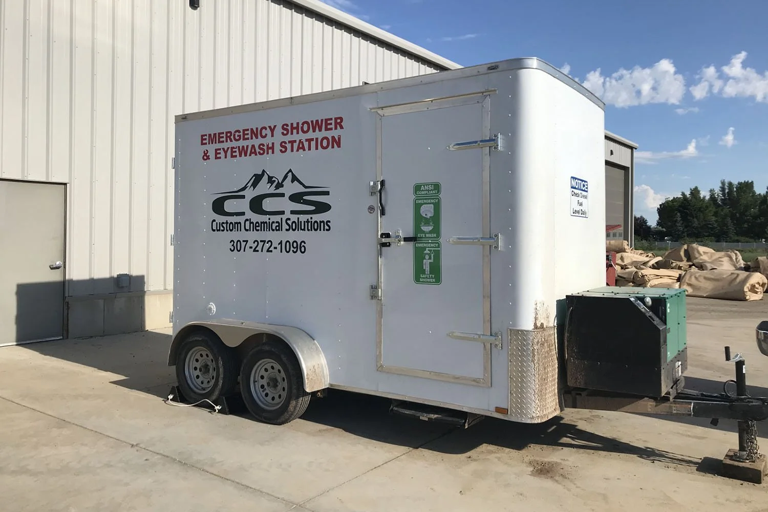 Trailer labeled 'Emergency Shower & Eyewash Station' with 'Custom Chemical Solutions' logo and contact number, parked outside a building with a closed door and a pile of bags in the background.