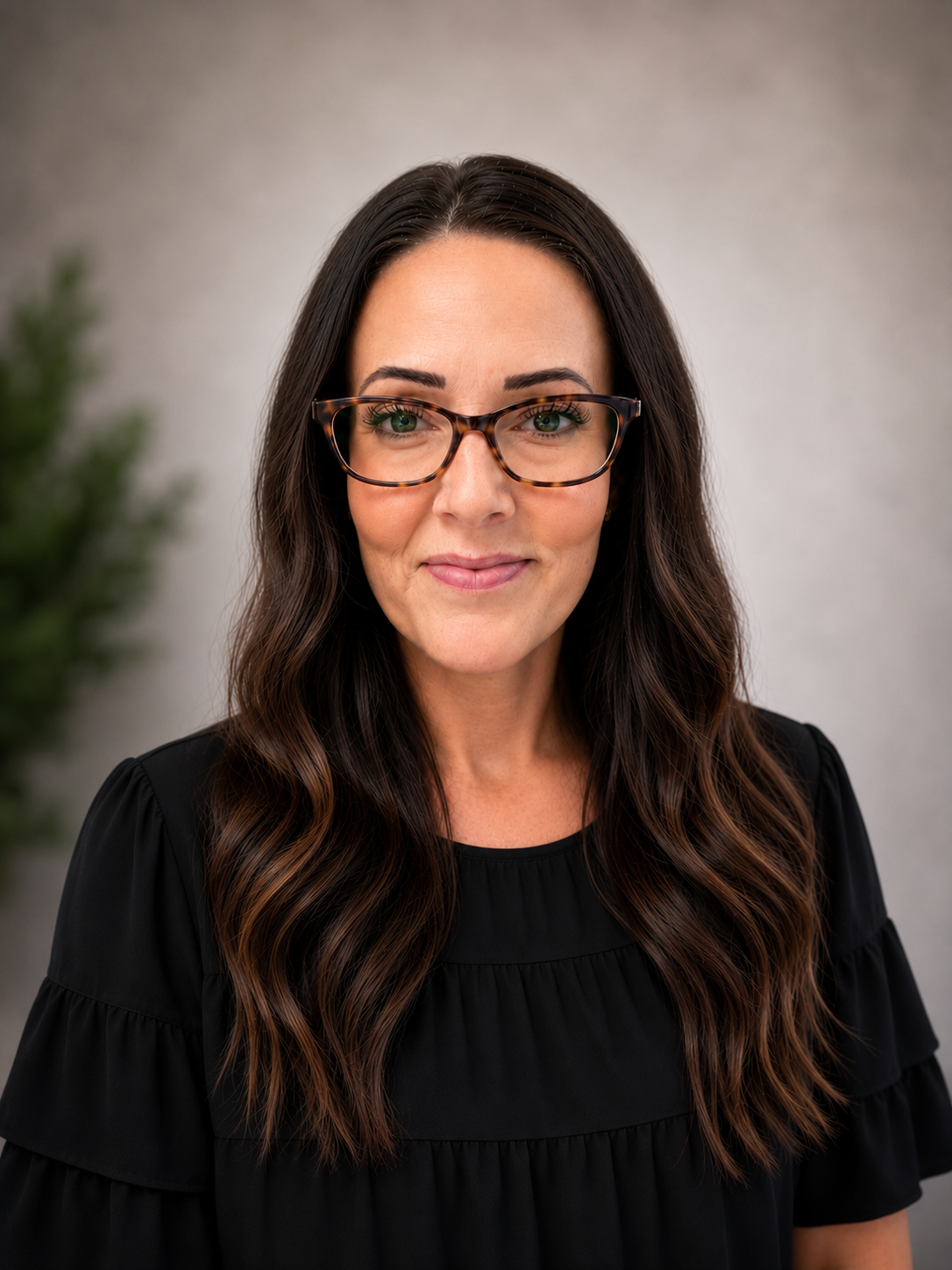 Close-up portrait of a woman with long wavy brown hair, wearing tortoiseshell glasses and a black top, smiling slightly, with a blurred green plant and gray background.