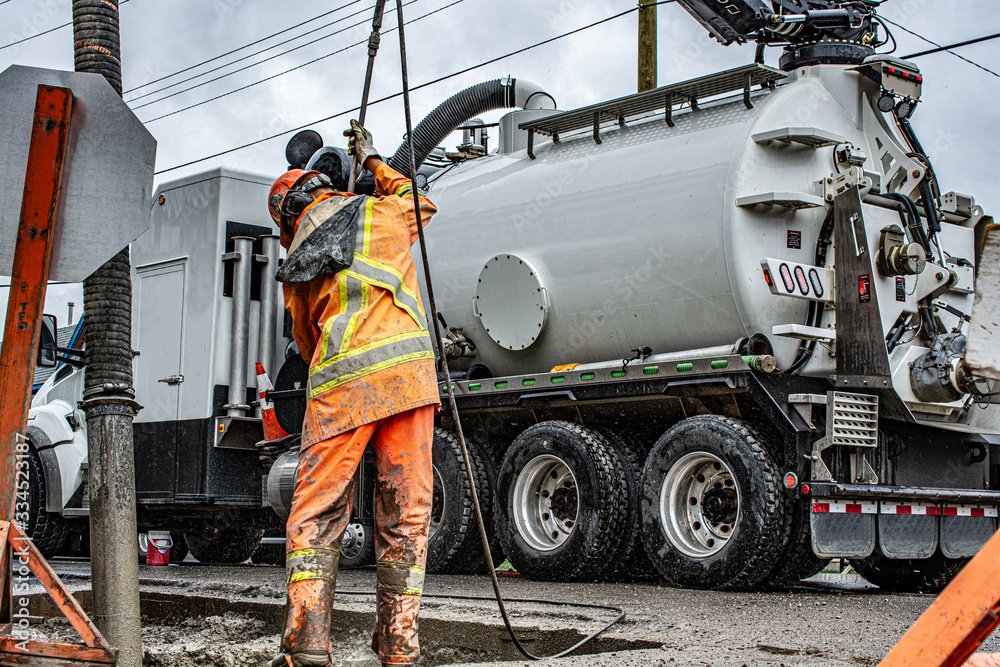A firefighter in orange and yellow protective gear rakes mud off a large vacuum truck on a wet street in rainy weather.