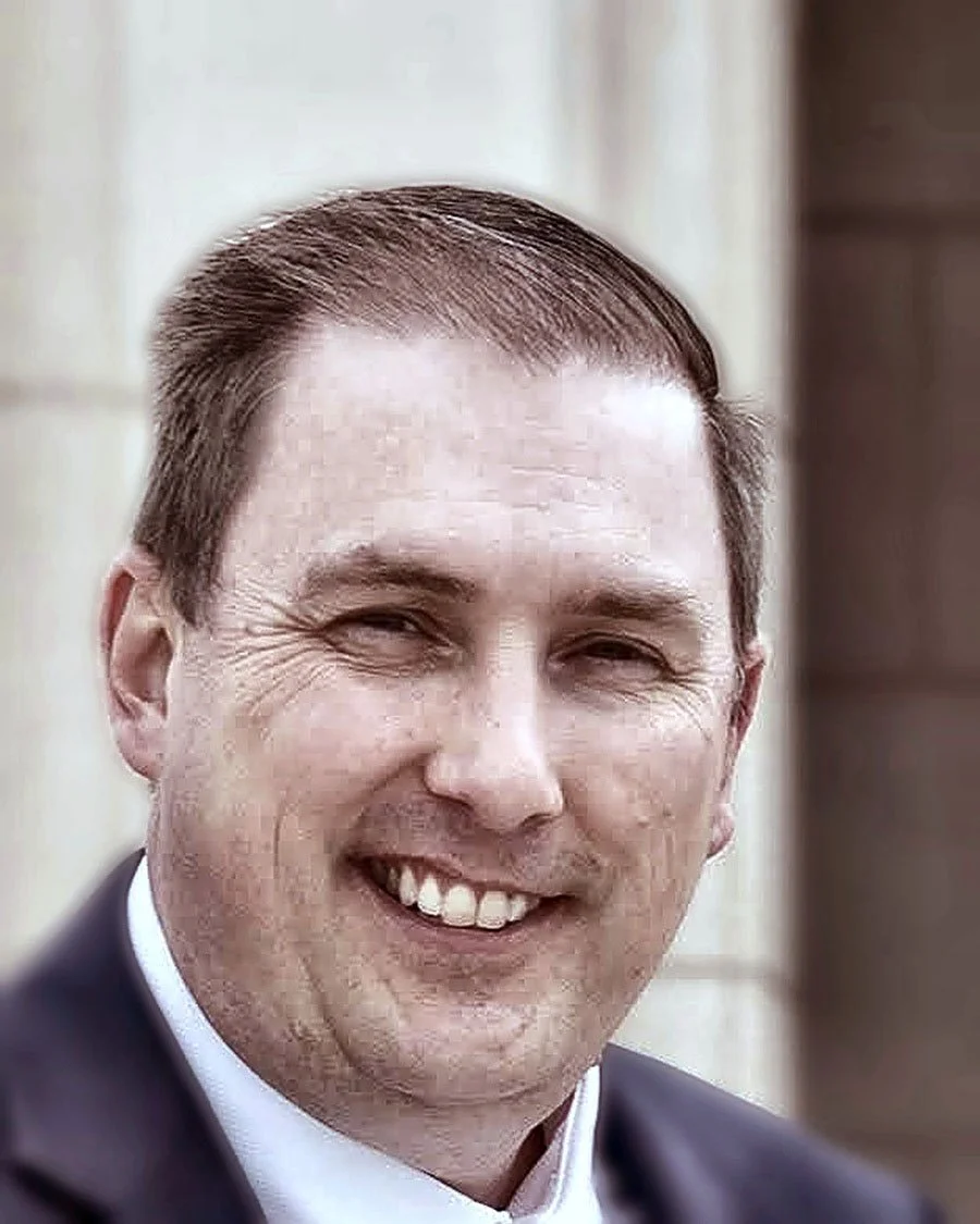 A smiling man with short, dark hair wearing a suit and tie, outdoors with a blurred building background.