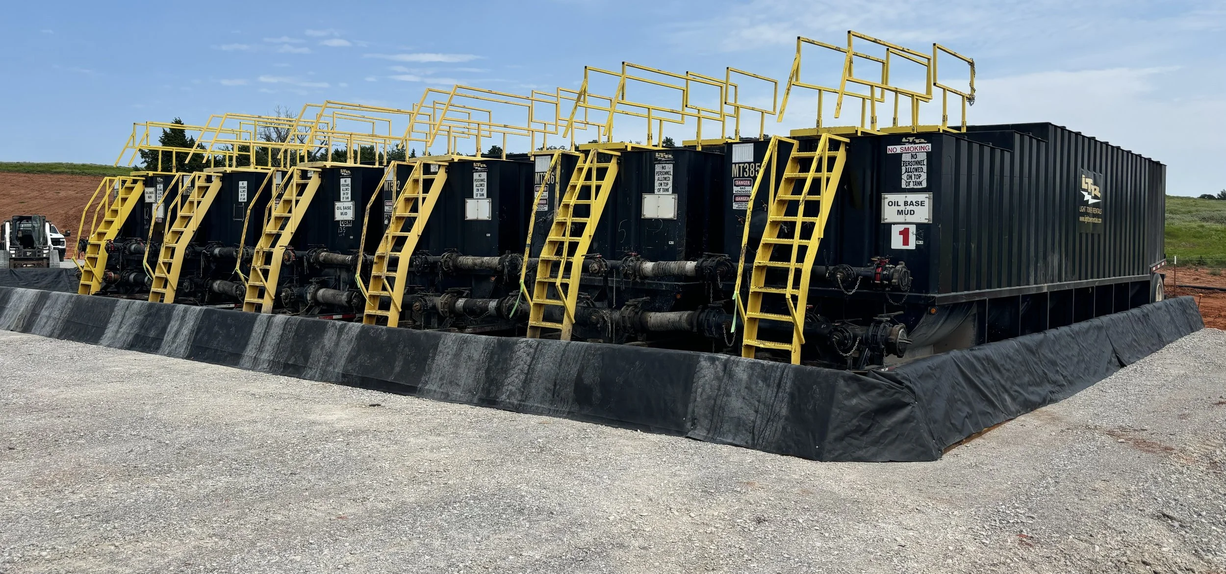 Large black oil containment tank on a construction site with yellow staircases and railings, and interconnected pipes at the base, surrounded by gravel and dirt, with a pickup truck and clear sky in the background.