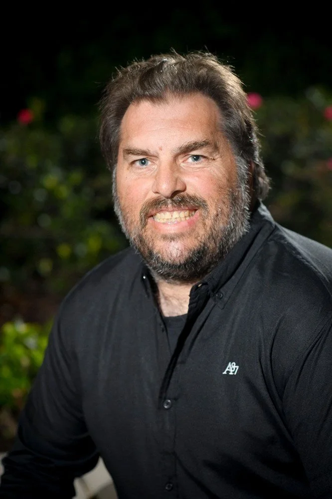 A man with a beard, wearing a black button-up shirt, smiling outdoors with a blurred green and pink floral background.