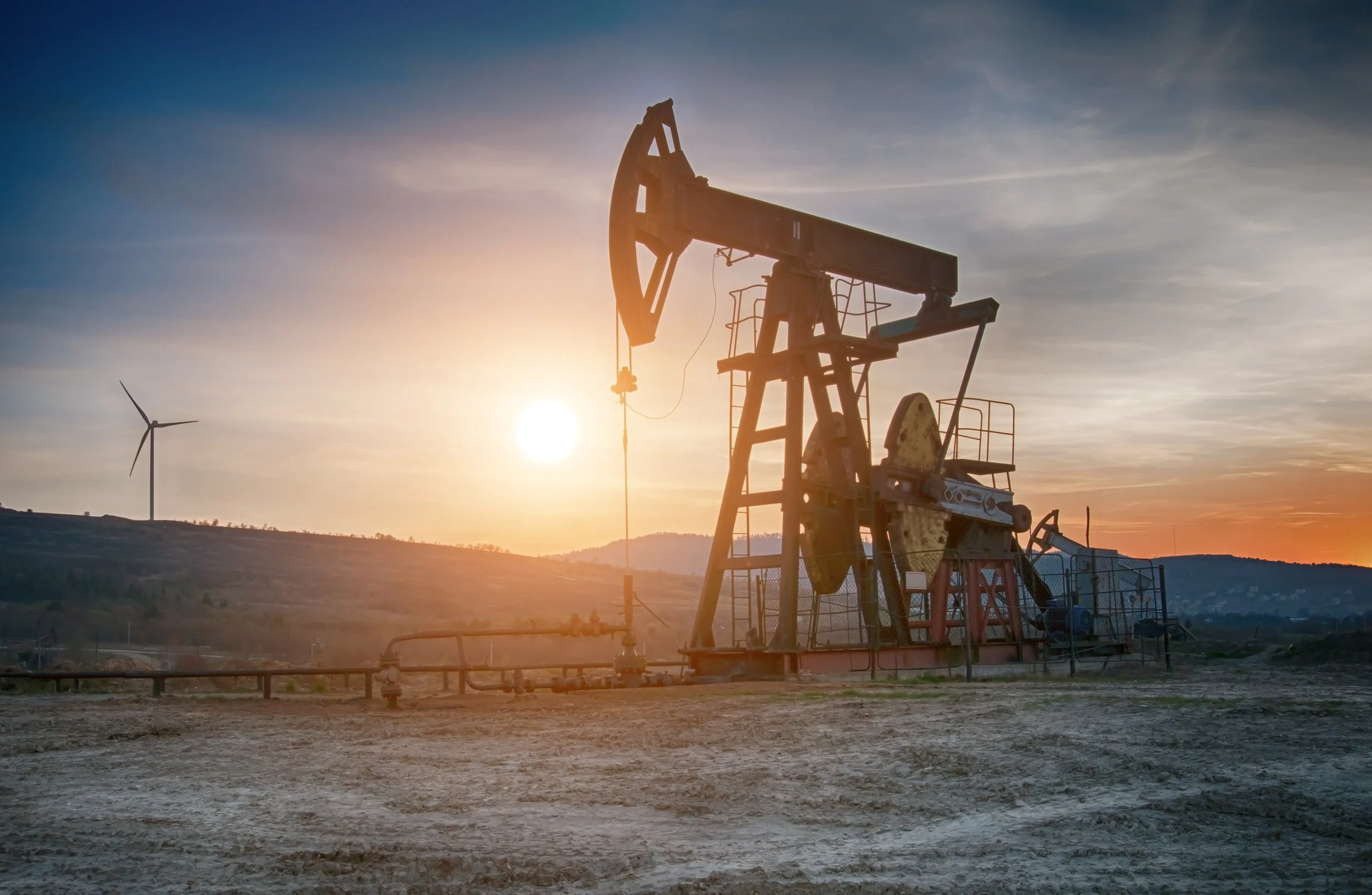 Oil pumpjack silhouette against sunset sky with wind turbine in the background
