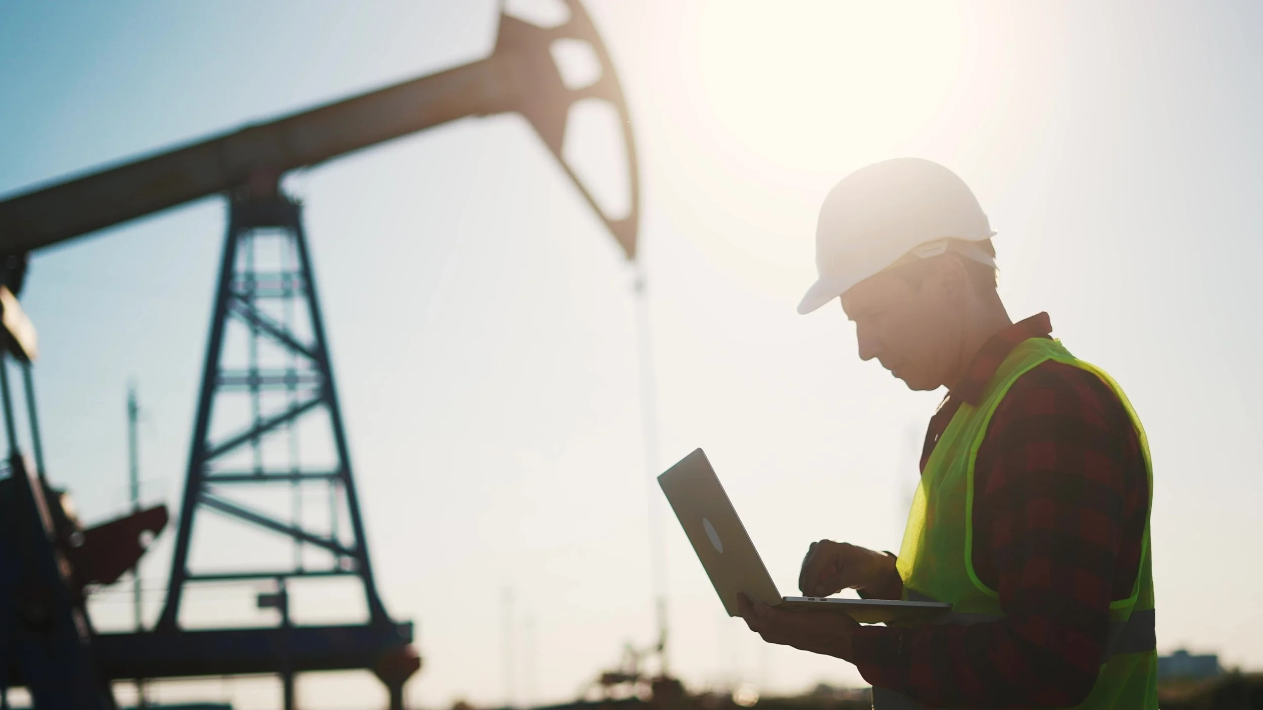 A construction worker wearing a hard hat and safety vest is working outdoors, holding a tablet or device, with an oil drilling rig in the background during the daytime.