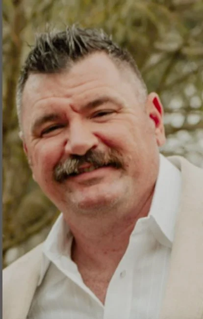 A smiling man with a mustache and short hair outdoors with trees in the background.