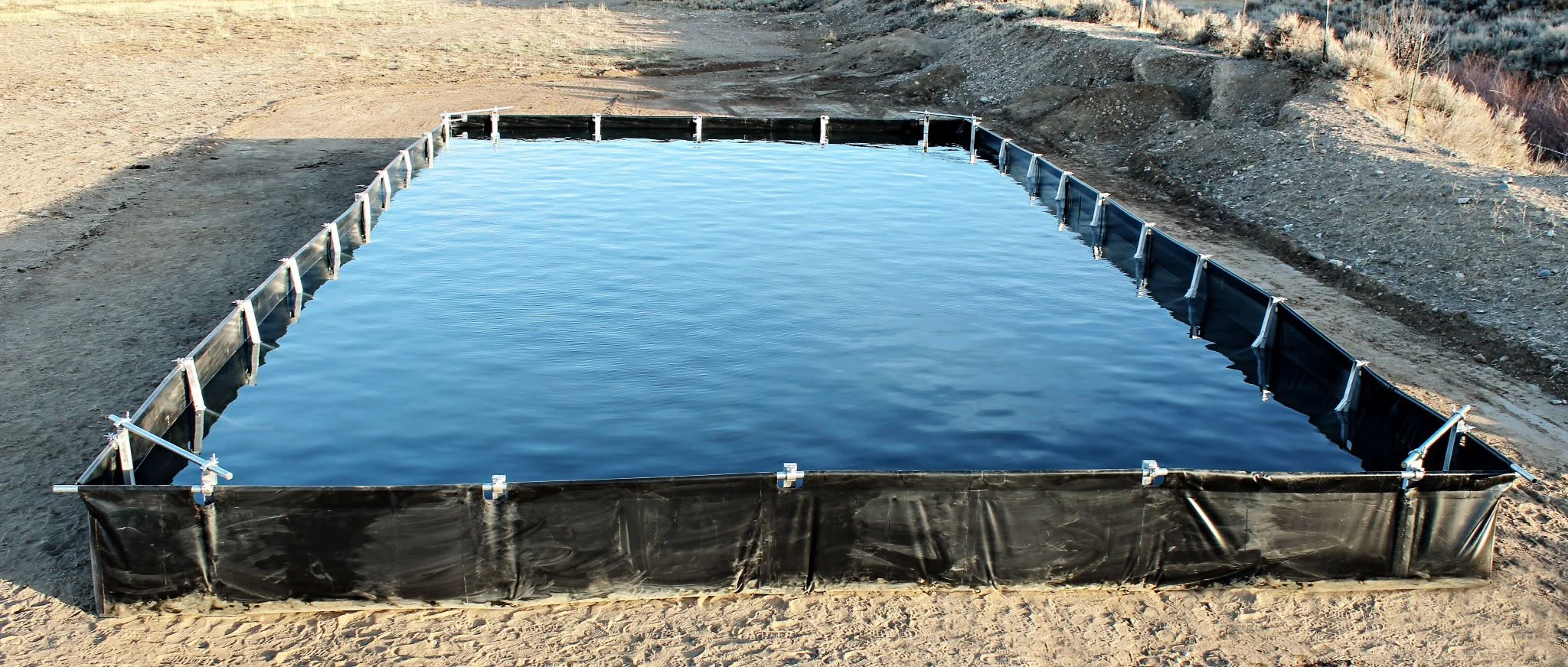 A rectangular water reservoir with a black liner, situated on a dirt road in a desert landscape with sparse vegetation.