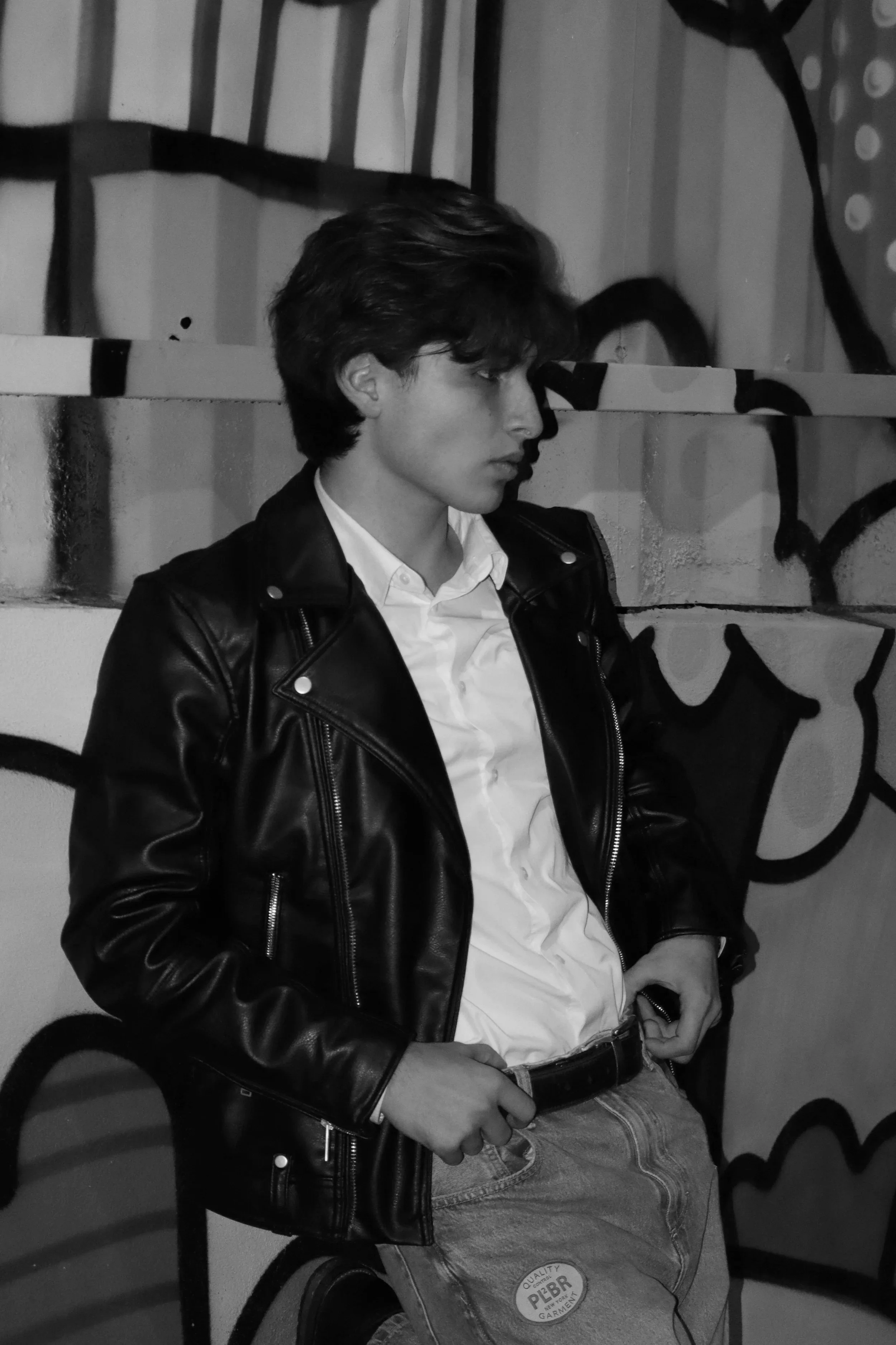 Un joven con cabello oscuro, usando una chaqueta de cuero y camisa blanca, sentado en una silla de plástico frente a una pared con graffiti en blanco y negro.
