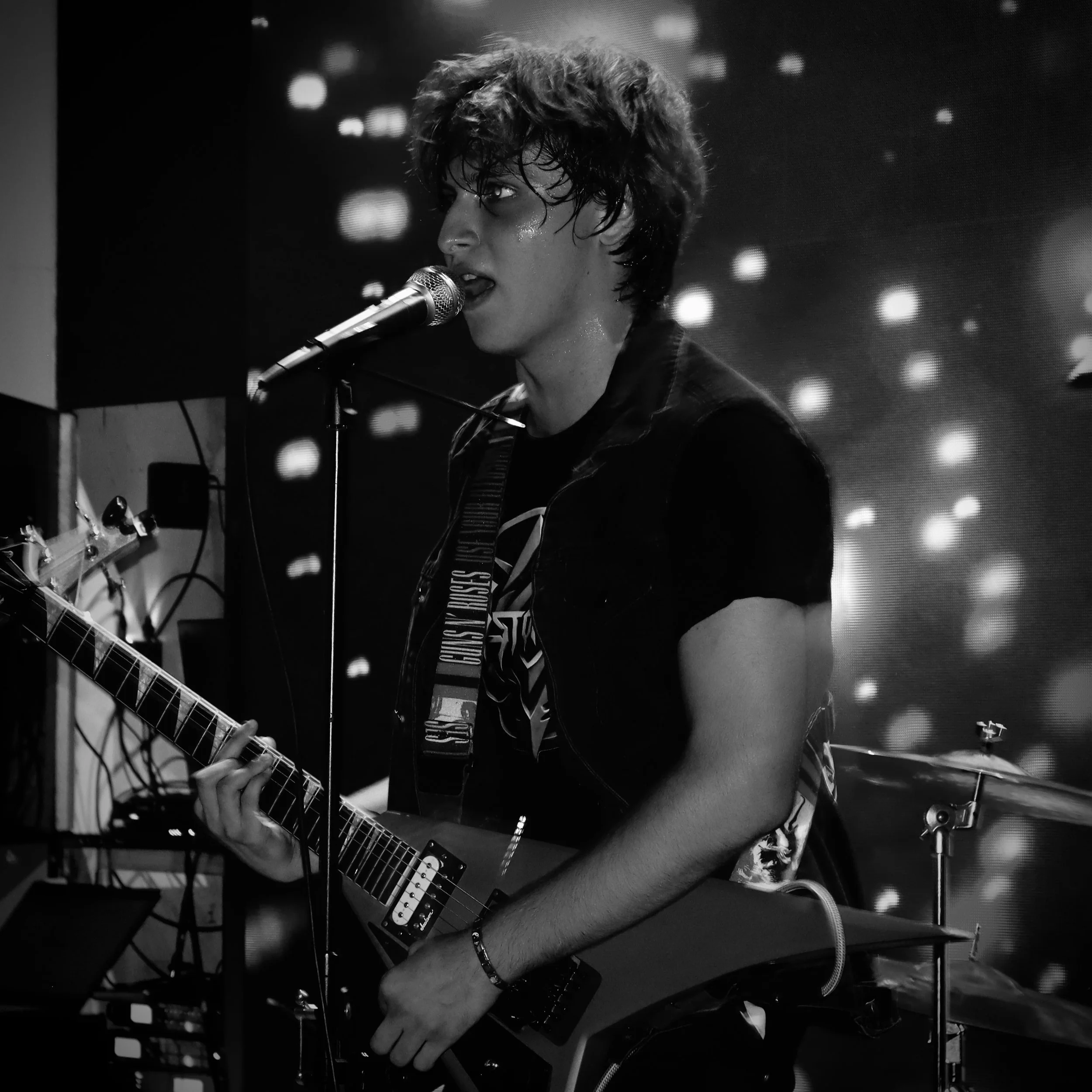 Joven músico tocando guitarra eléctrica y cantando en un escenario, con micrófono, en un ambiente oscuro con luces de fondo difusas.
