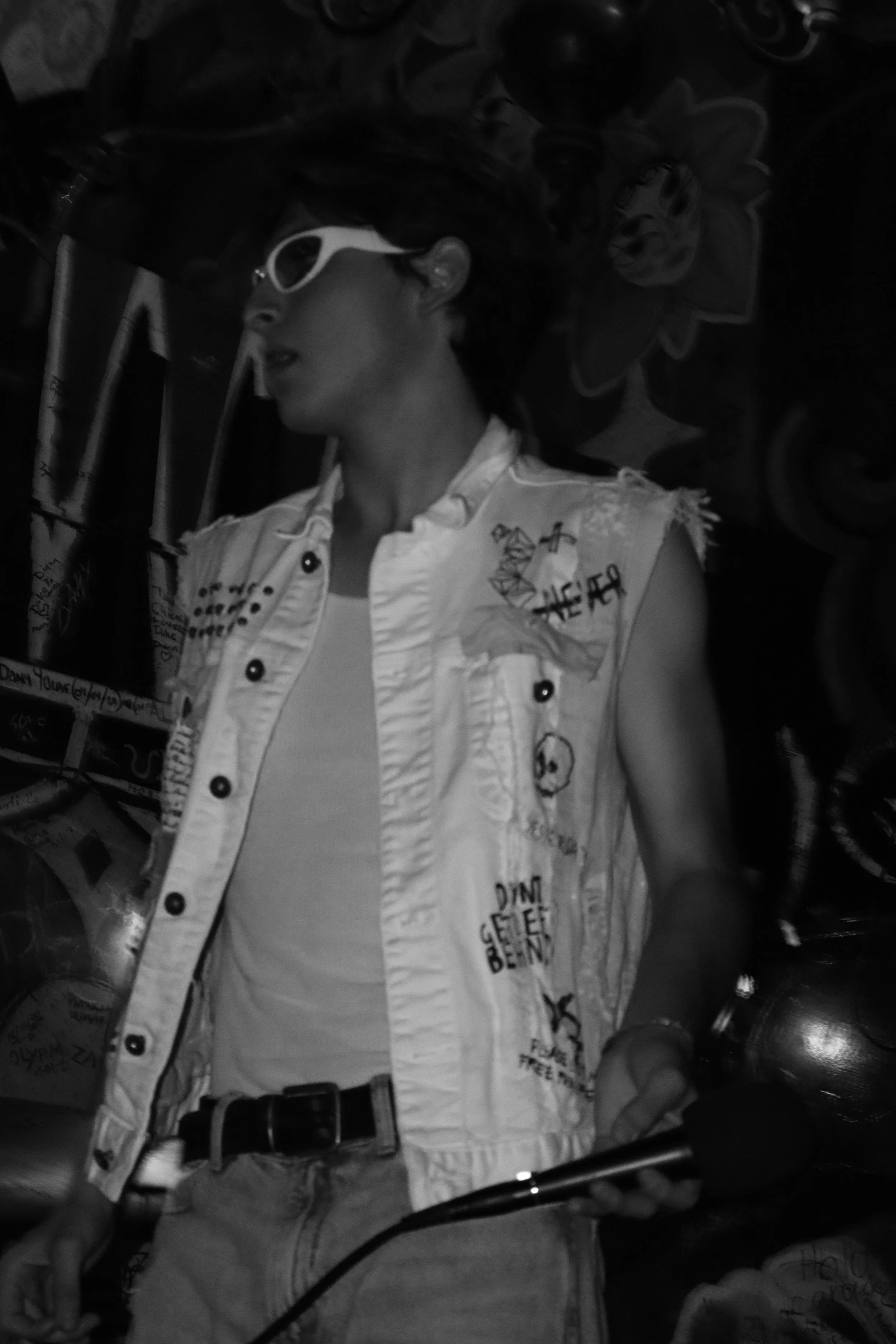Joven con cabello rizado, gafas blancas y camiseta sin mangas, en un entorno oscuro con decoraciones de flores.
