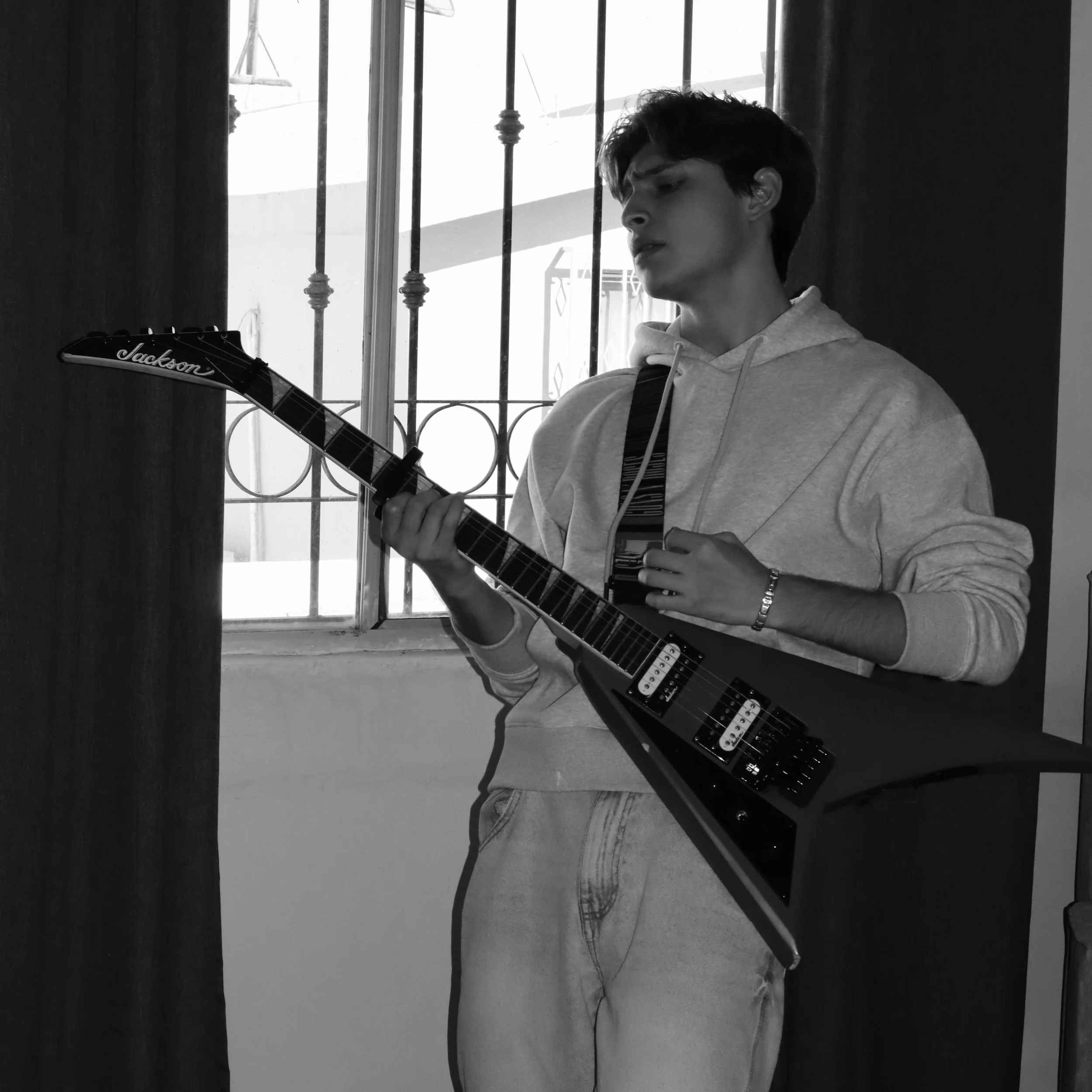 Joven tocando una guitarra eléctrica en un interior junto a una ventana con rejas.