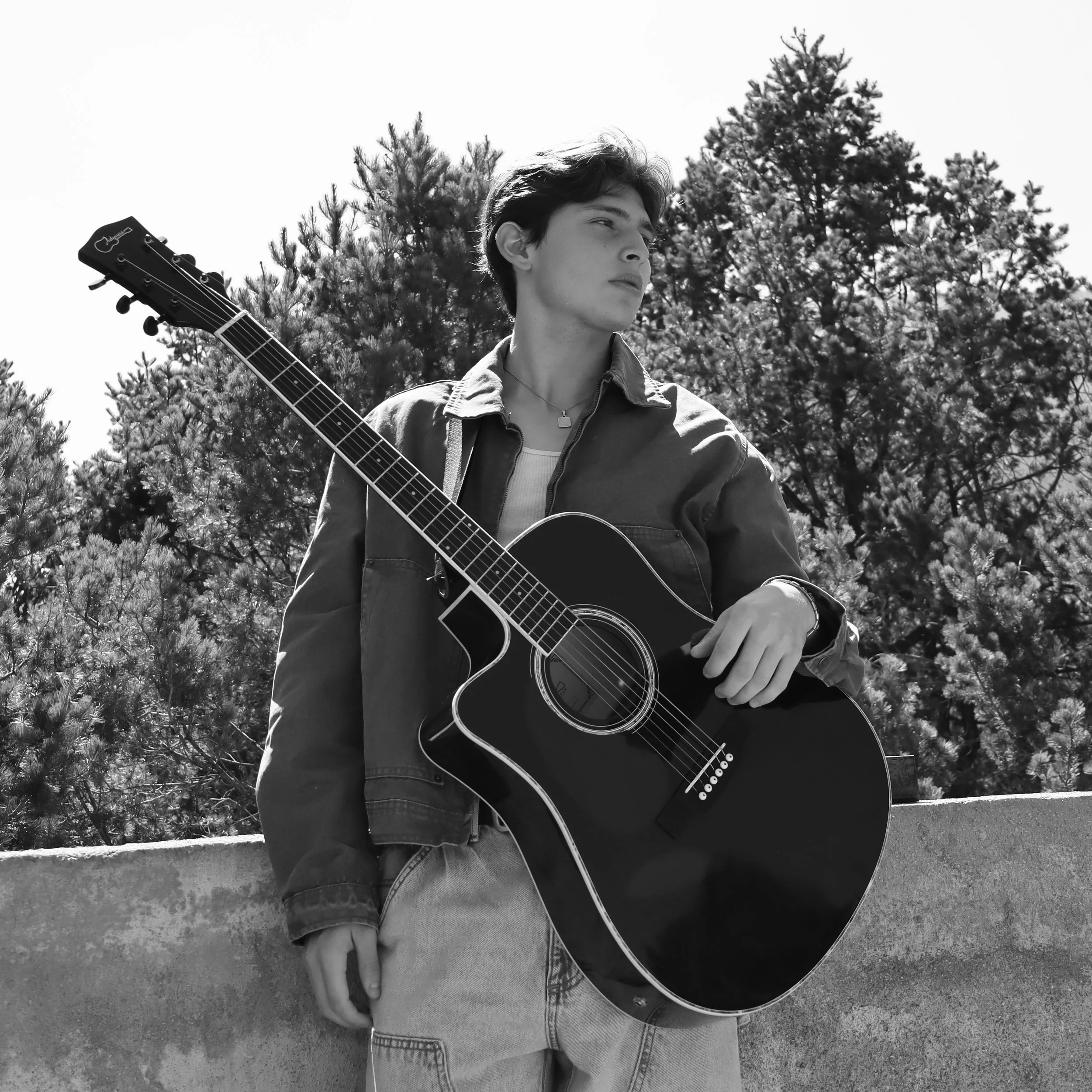 Joven tocando guitarra acústica en un espacio al aire libre con árboles de fondo en blanco y negro.