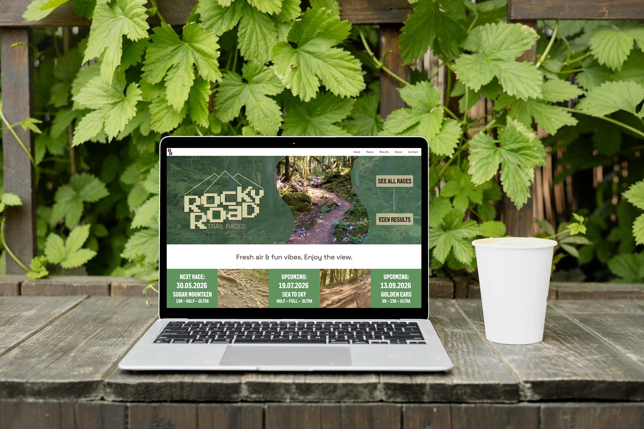 A laptop mockup of a trail race website, placed on a wooden table outdoors, with lush green leaves and a paper cup nearby.