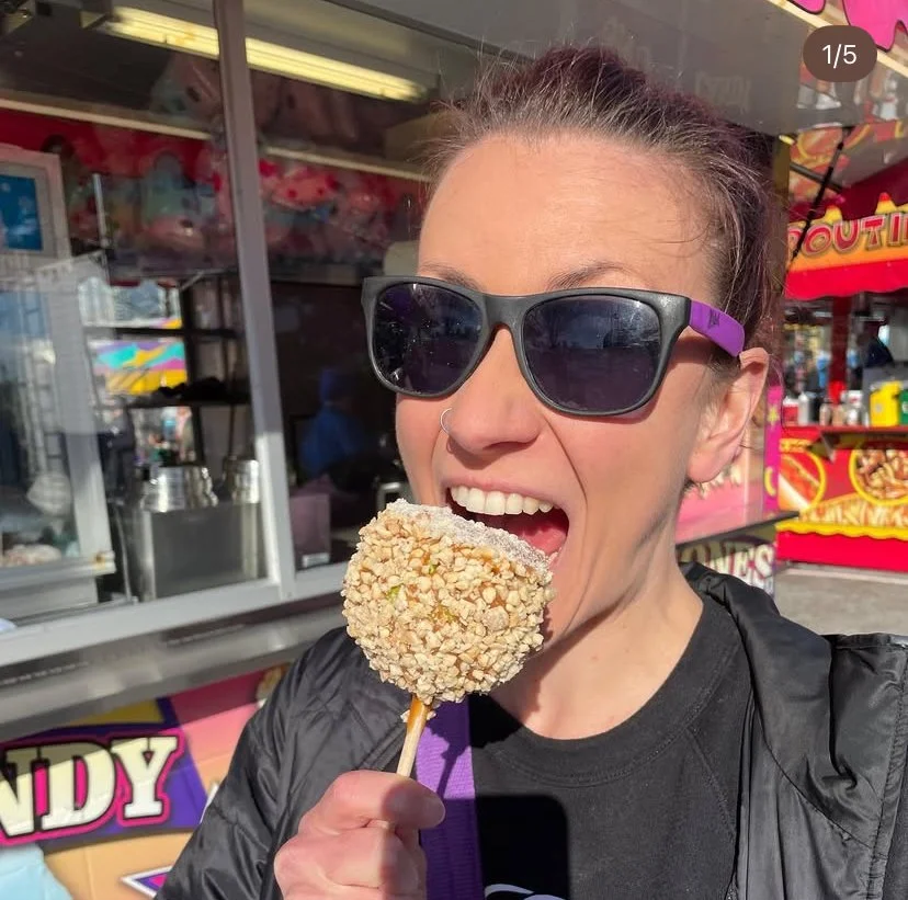 Lisa wearing sunglasses and a black jacket, holding and eating a caramel apple coated with chopped nuts at a carnival.