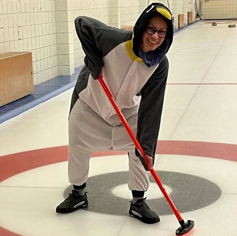 Lisa, dressed as a penguin, smiling and holding a curling broom on an indoor curling rink.