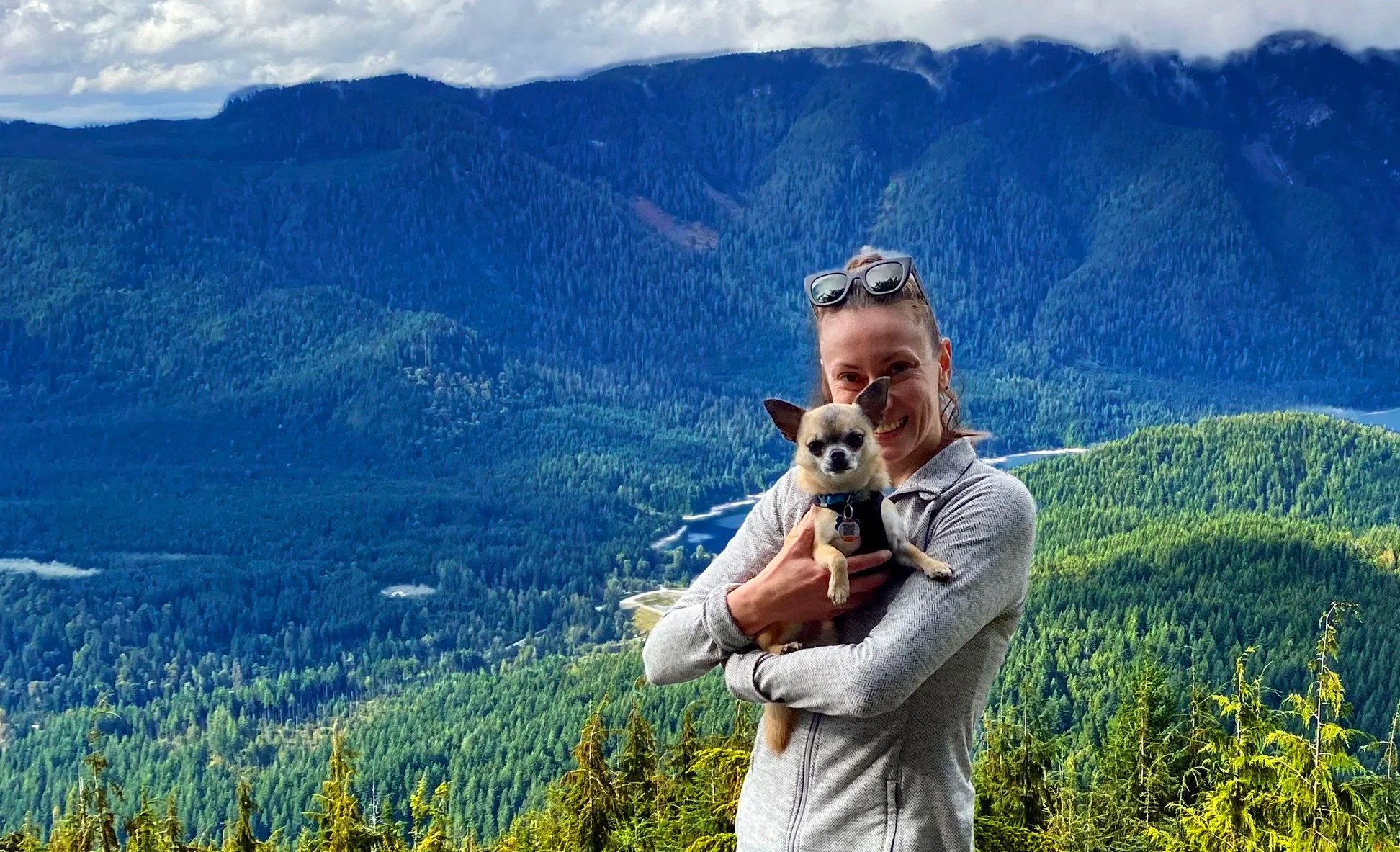 A photo of Lisa holding her chihuahua Arthur, standing outdoors on a hillside with a view of a forested mountain landscape in the background. Lisa is smiling and wearing sunglasses on her head.