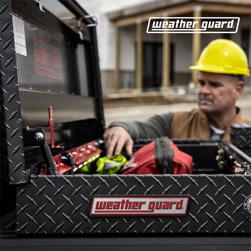 A man wearing a yellow safety helmet inspecting tools and equipment in a black Weather Guard toolbox at a construction site.