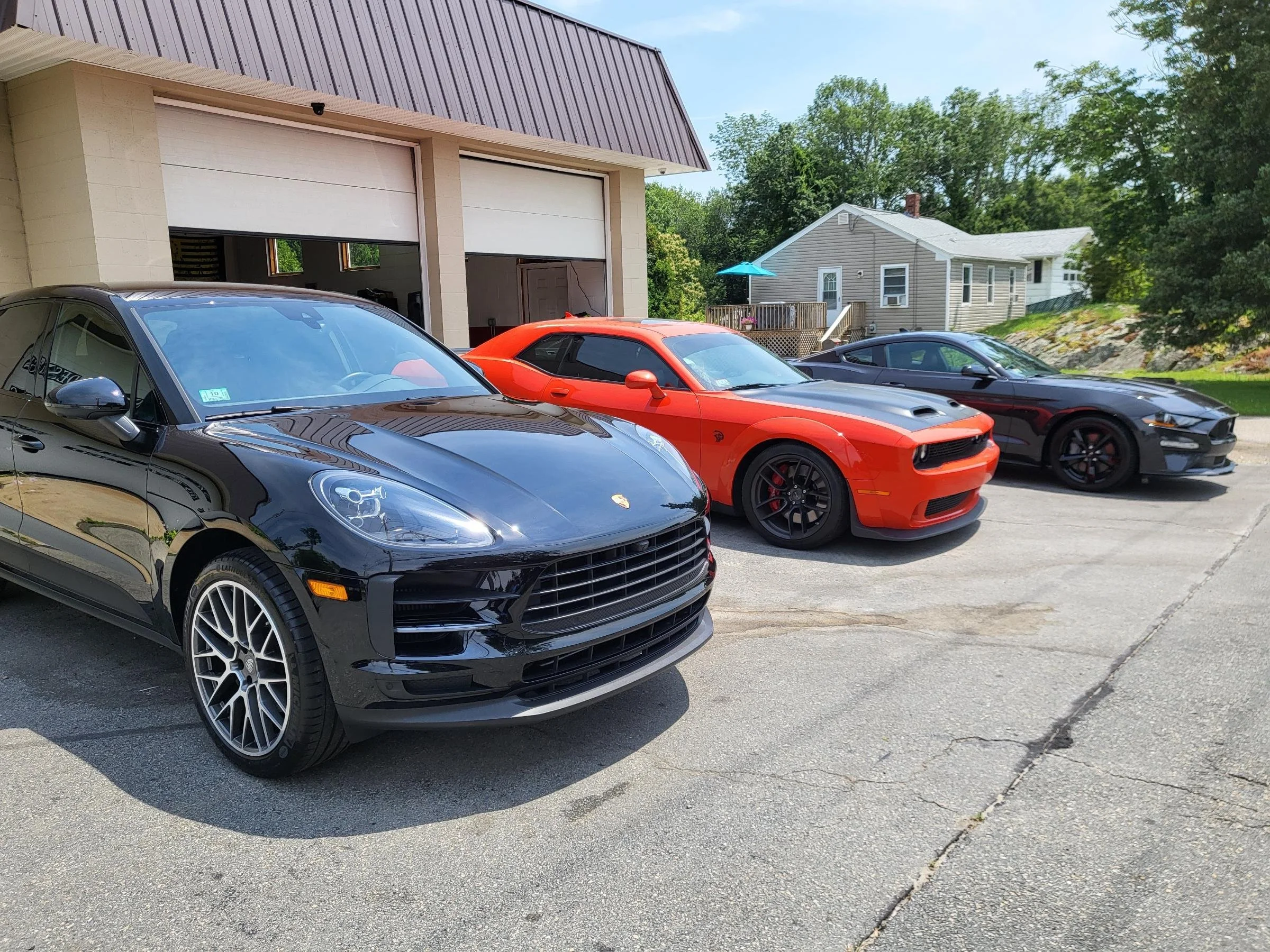 Three luxury cars, a black Porsche, a red Dodge Challenger, and a black Chevrolet Camaro, parked in front of a garage with a beige house and green trees in the background.