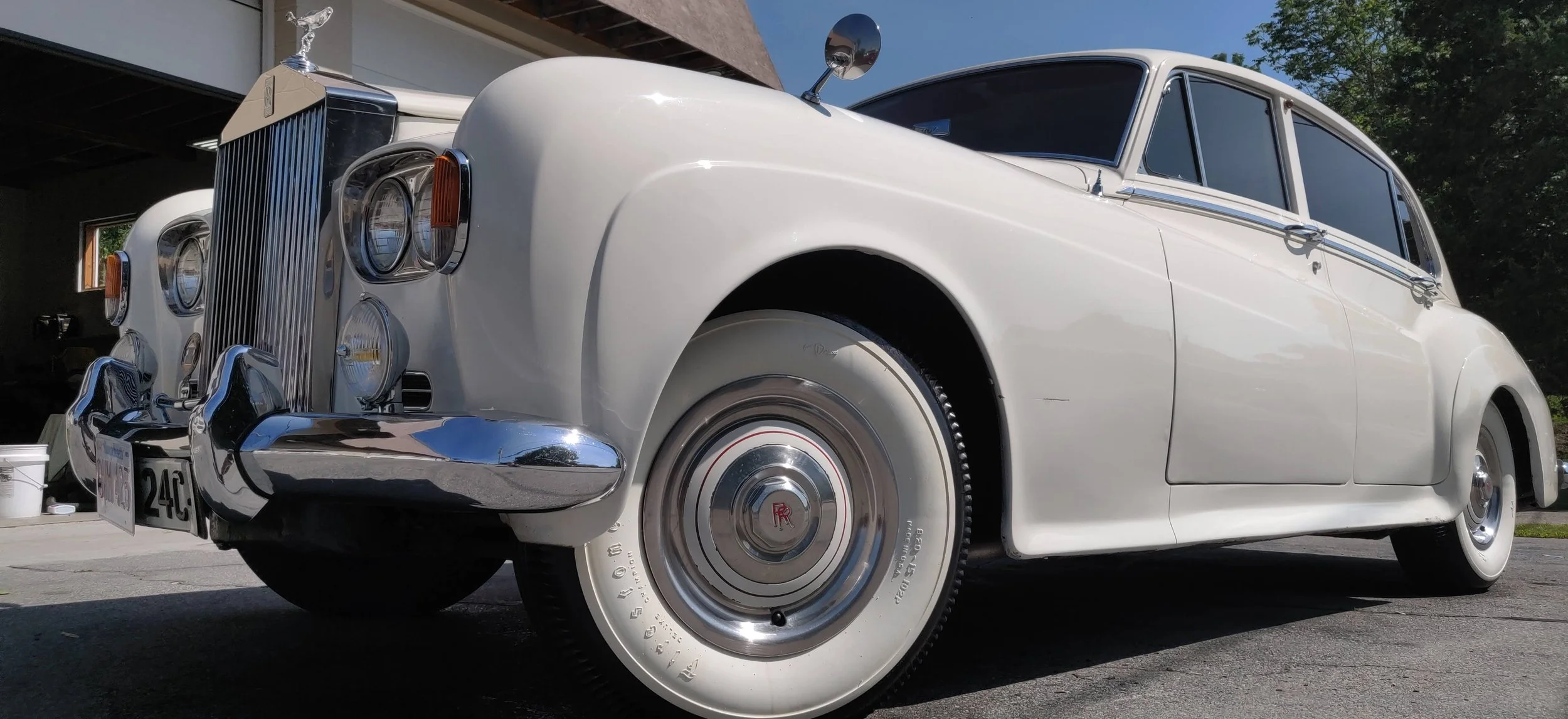 A vintage white Rolls-Royce car parked outside, showcasing its classic front grille, round headlights, chrome bumper, and whitewall tires.