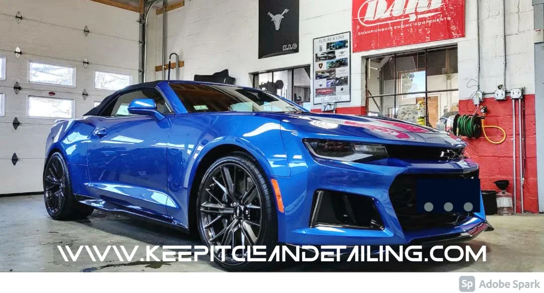 A blue Chevrolet Camaro convertible car parked inside a garage with various automotive posters and equipment visible in the background.