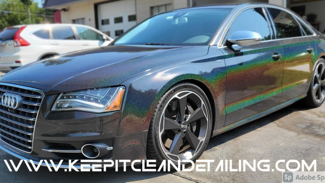 A black luxury sedan with a rainbow-colored iridescent wrap. Parked outside a building, with other vehicles nearby.