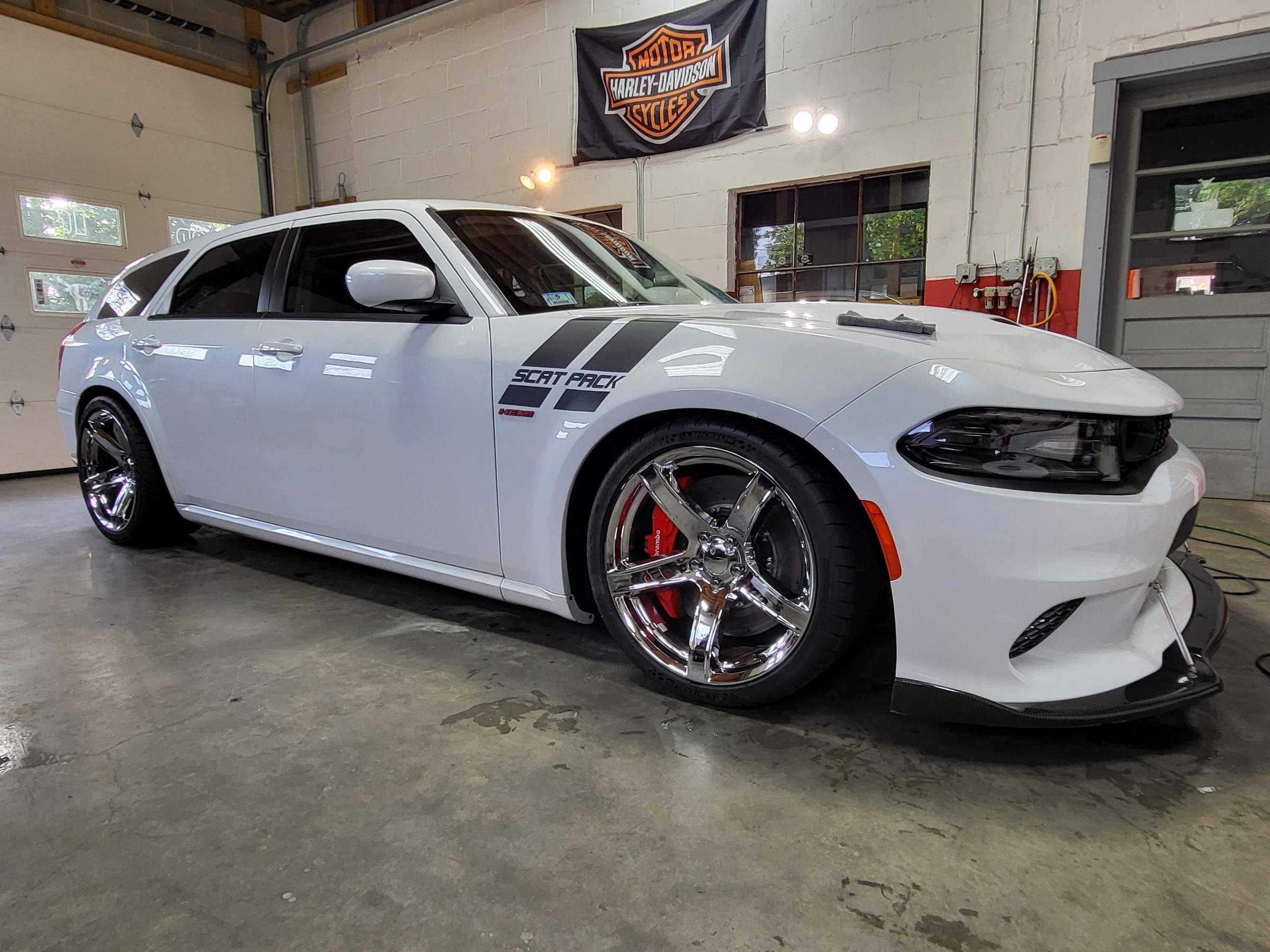A white sports car parked inside a garage with a Harley-Davidson flag hanging on the wall. The car has black decals and chrome wheels, with a clean and shiny appearance.