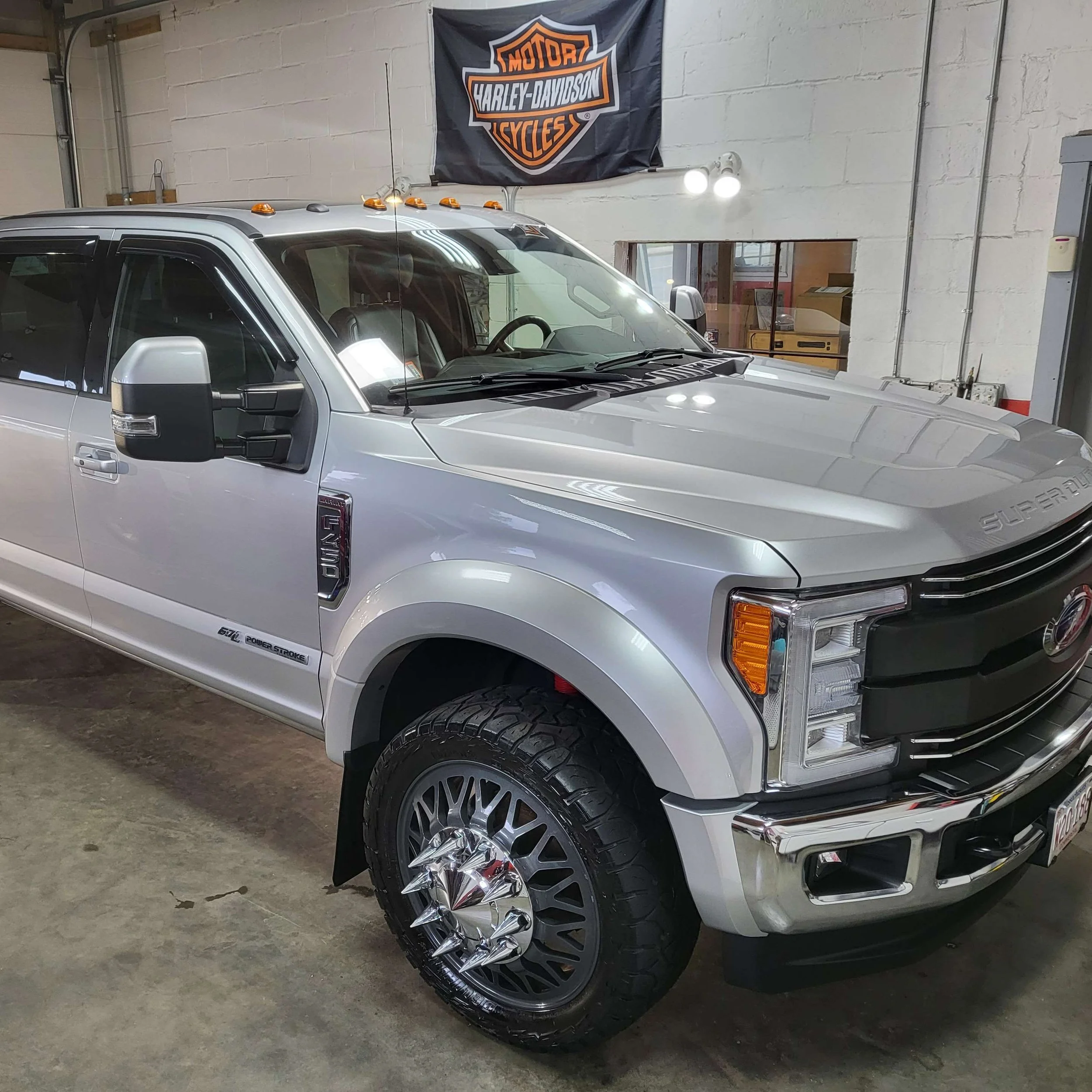 A silver pickup truck with black accents parked indoors in front of a wall with a Harley-Davidson flag. The truck has black wheels and chrome accents on the front grille and side mirrors. The front right wheel is visible, and the vehicle has characte