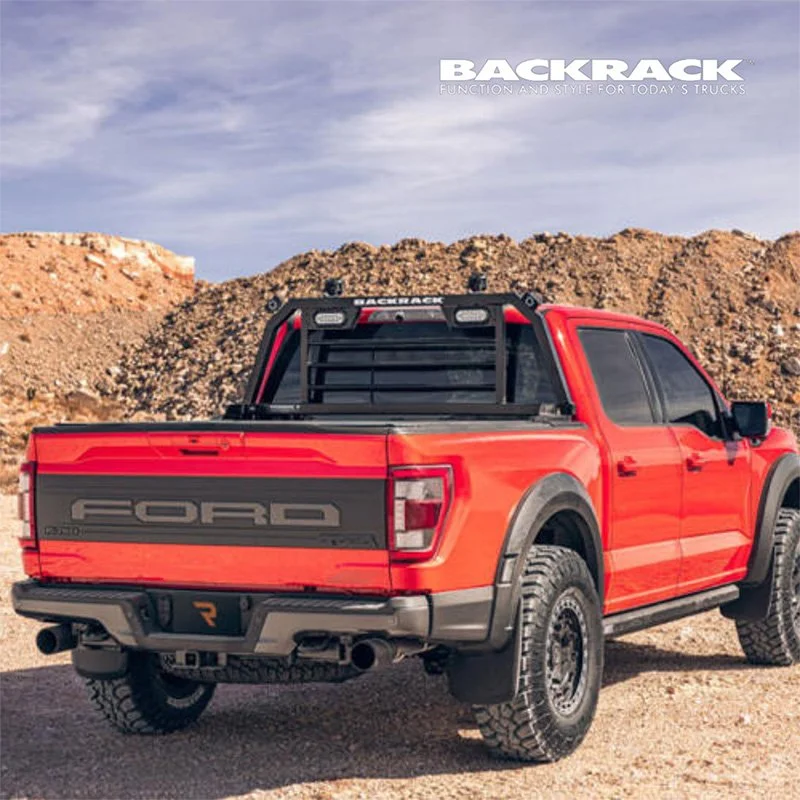 A red Ford pickup truck parked on dirt ground with rocky hills in the background, featuring a black custom backrack for truck bed storage and dual exhaust pipes.