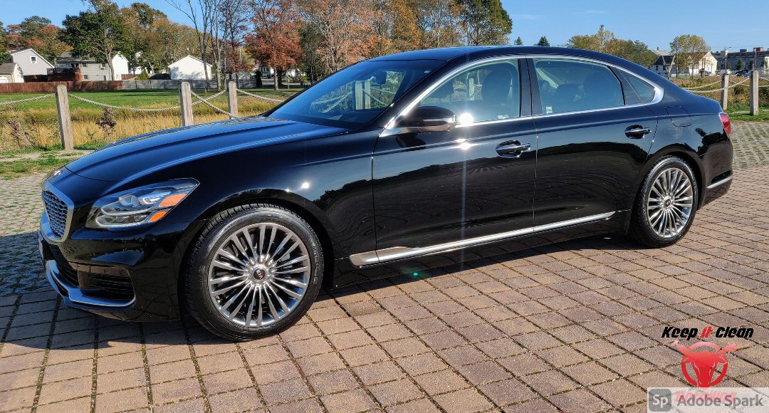 A black sedan parked on a brick-paved surface with a grassy area and trees in the background under a clear blue sky.