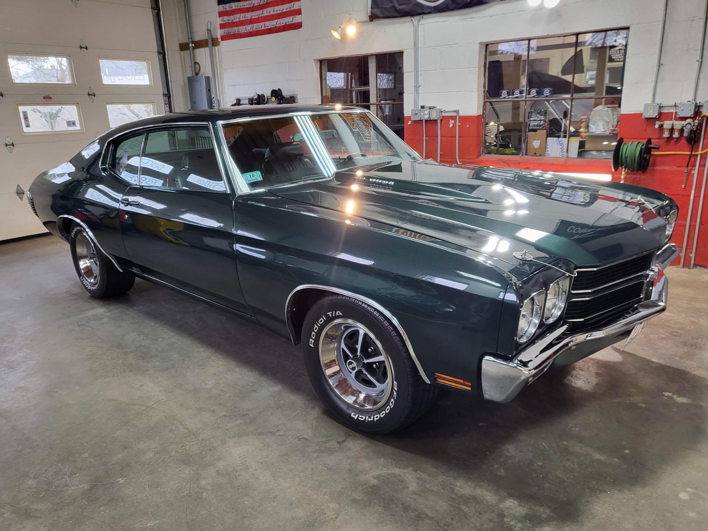 A black vintage muscle car parked inside a garage with a white wall and red stripes, metal pipes, and tools on the wall.
