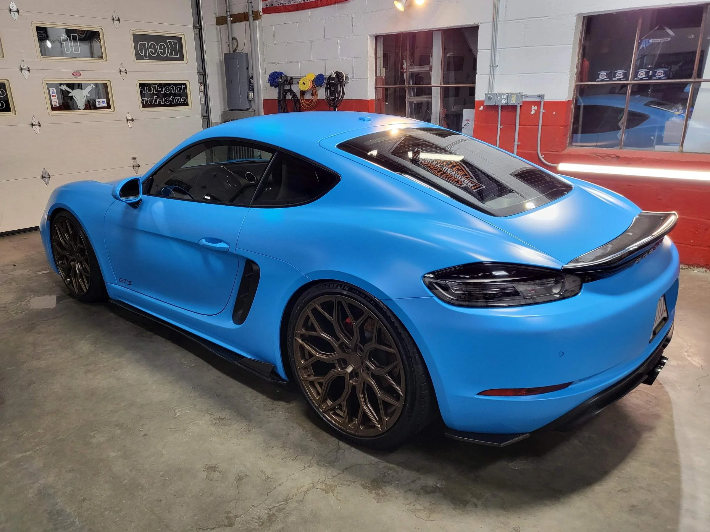 A bright blue sports car, likely a Porsche, inside a garage or workshop, with black wheels, parked near a garage door and behind a window showing a reflection of another blue vehicle.