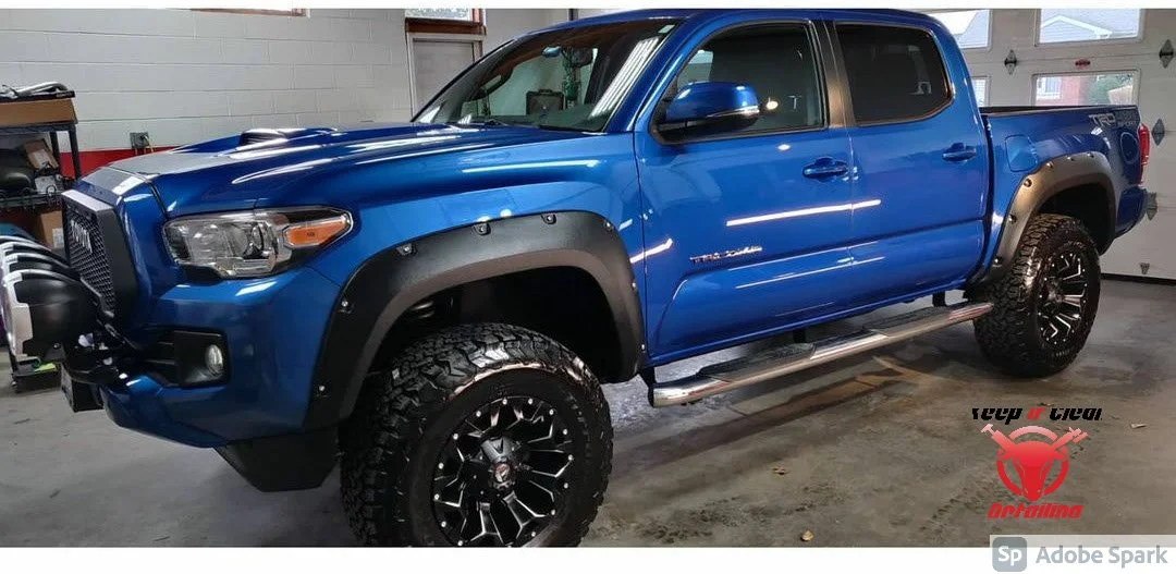 A blue Toyota Tacoma pickup truck with off-road modifications, including large tires, black fender flares, side steps, and a front bumper guard, parked inside a garage.