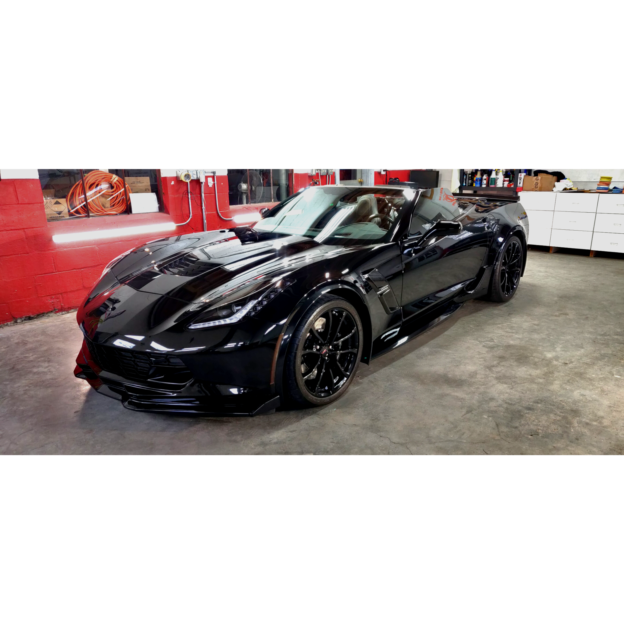 A black sports car parked in a garage with red walls, tools, and shelves in the background.