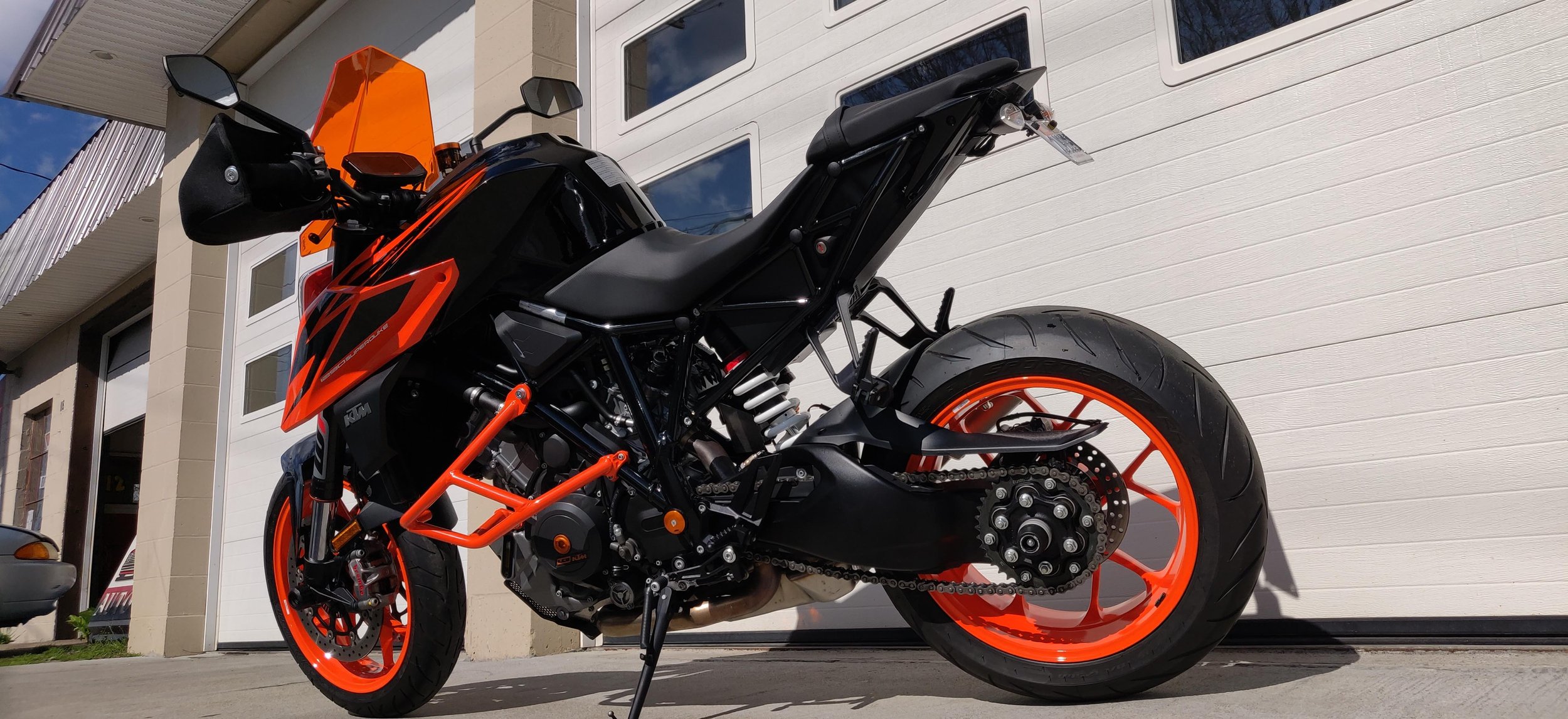 A black and orange KTM motorcycle parked outside a white garage with windows, against the background of a beige house with vertical siding and a blue sky with some clouds.