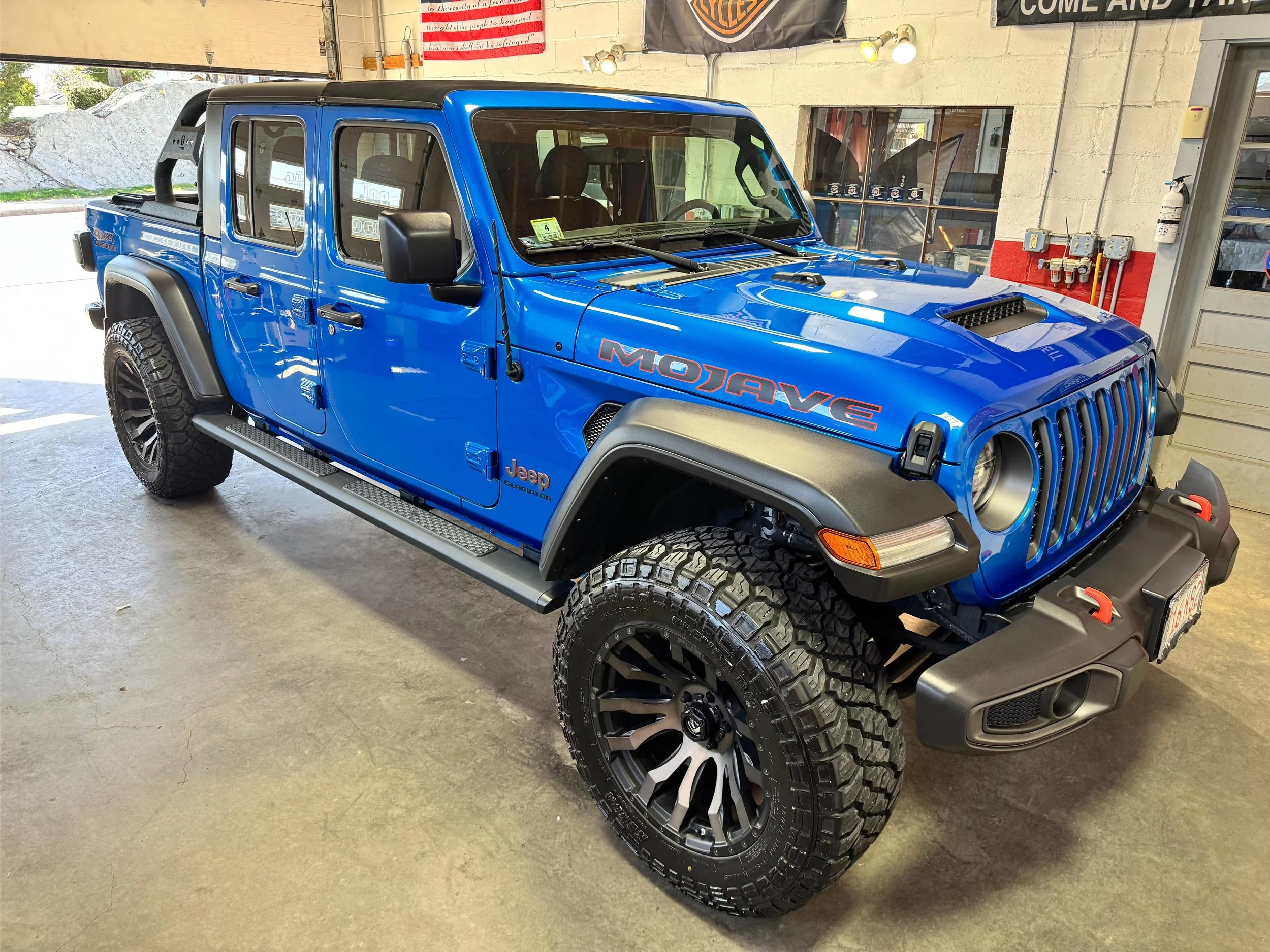 A blue Jeep Gladiator pickup truck with large off-road tires parked inside a garage. The garage has a flag and banners on the wall and various electrical and safety equipment.