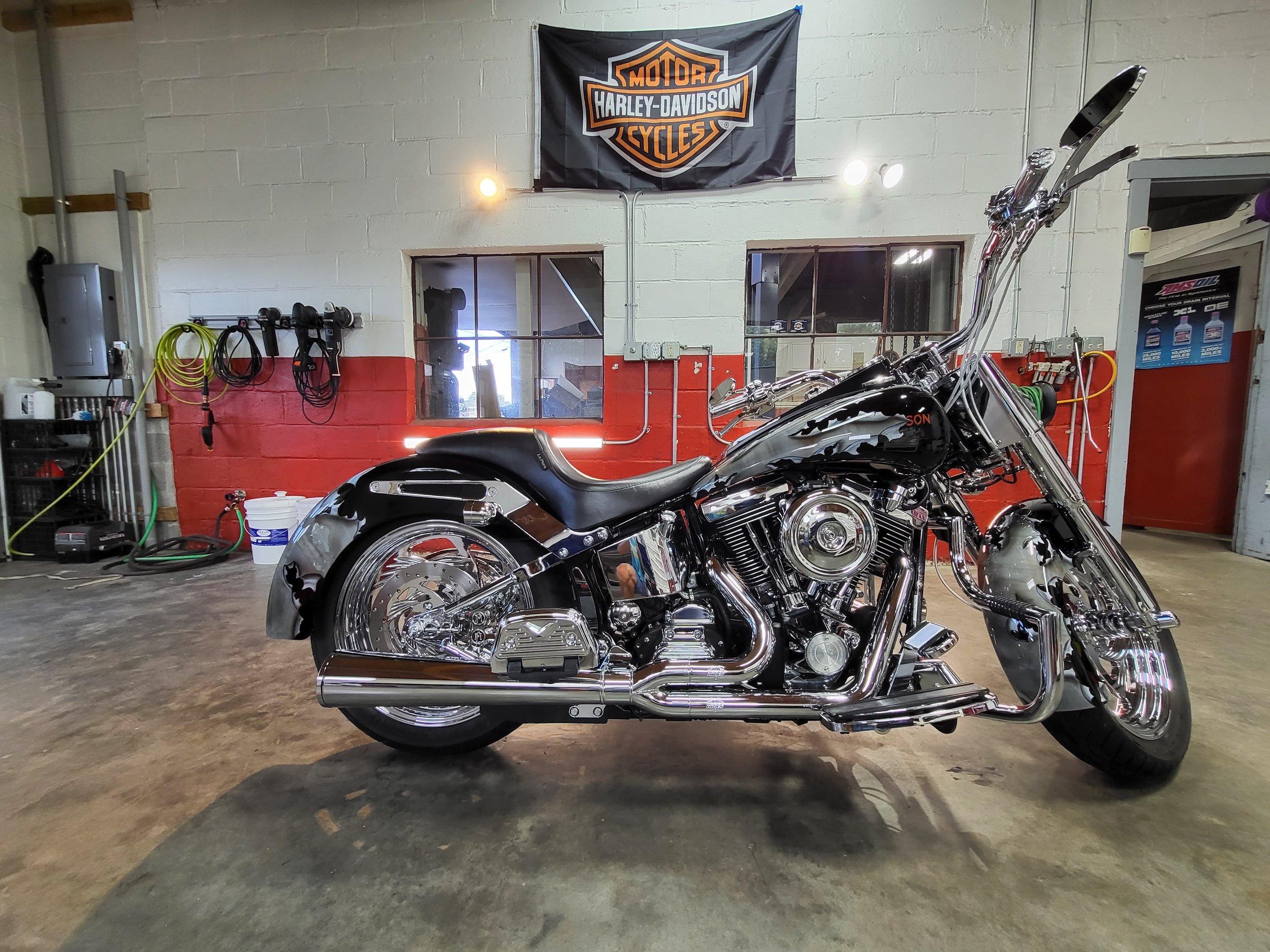 A black Harley-Davidson motorcycle is parked inside a garage with a Harley-Davidson banner on the wall. The garage has tools and equipment along the wall.