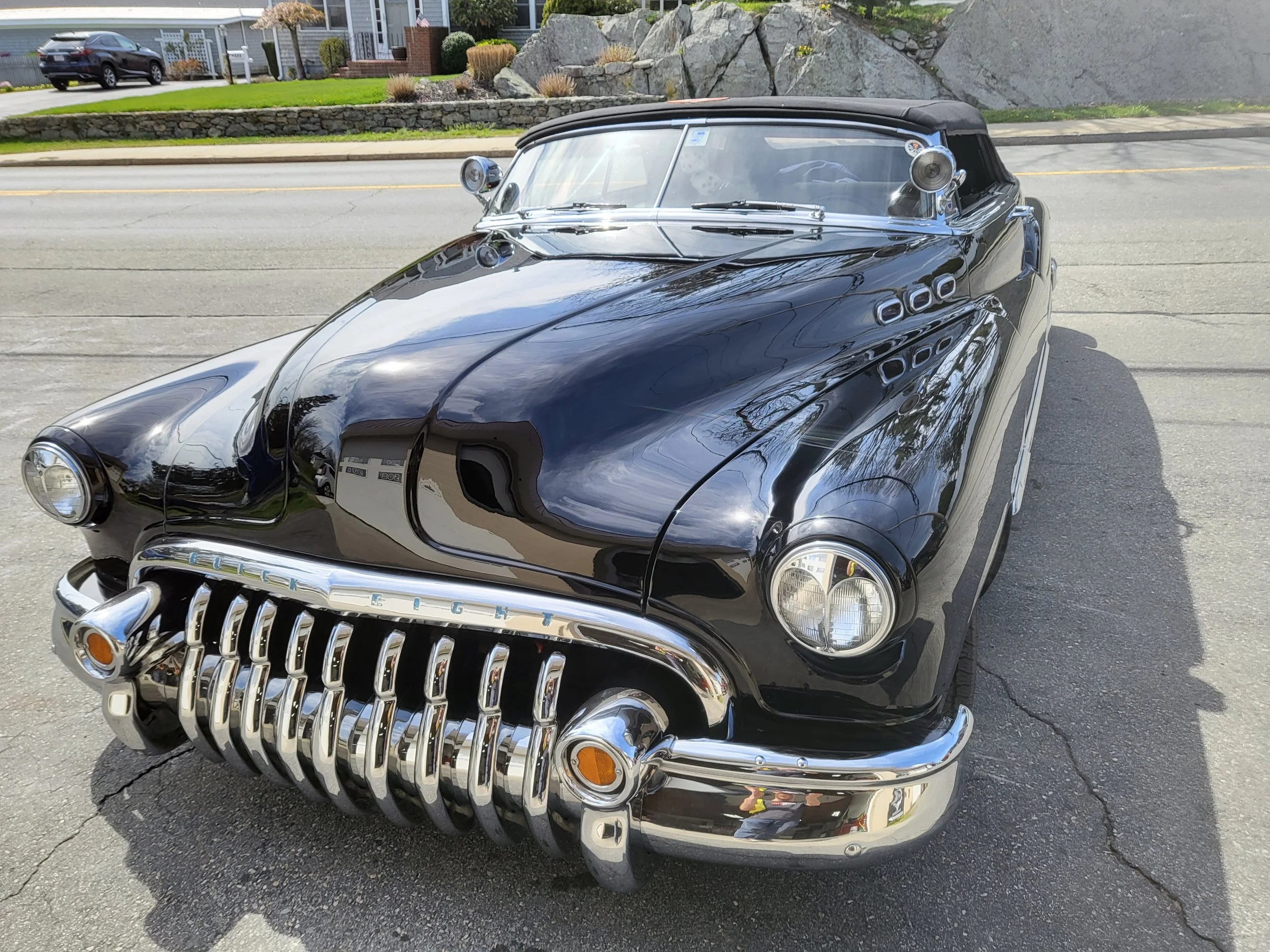 A shiny black vintage car with chrome details parked on a street in front of a house with a lawn and rocky garden.