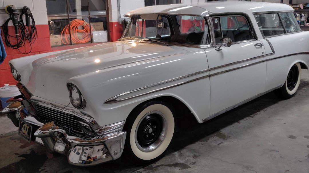 A vintage light gray station wagon with white-wall tires parked inside a garage.