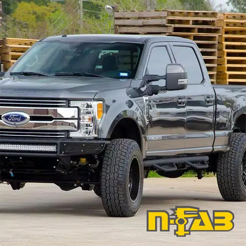 A gray Ford pickup truck with large off-road tires, lifted suspension, and aftermarket front bumper parked on a concrete surface with stacked wood pallets in the background.
