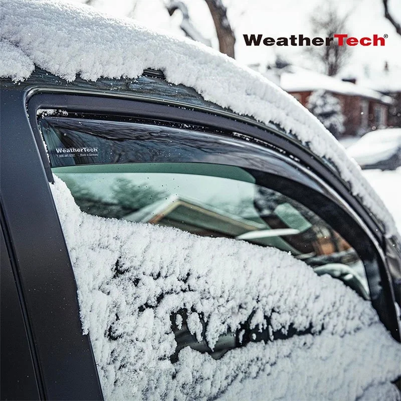 A car with snow on its windows and roof, parked outdoors in a snowy neighborhood with houses and trees in the background.