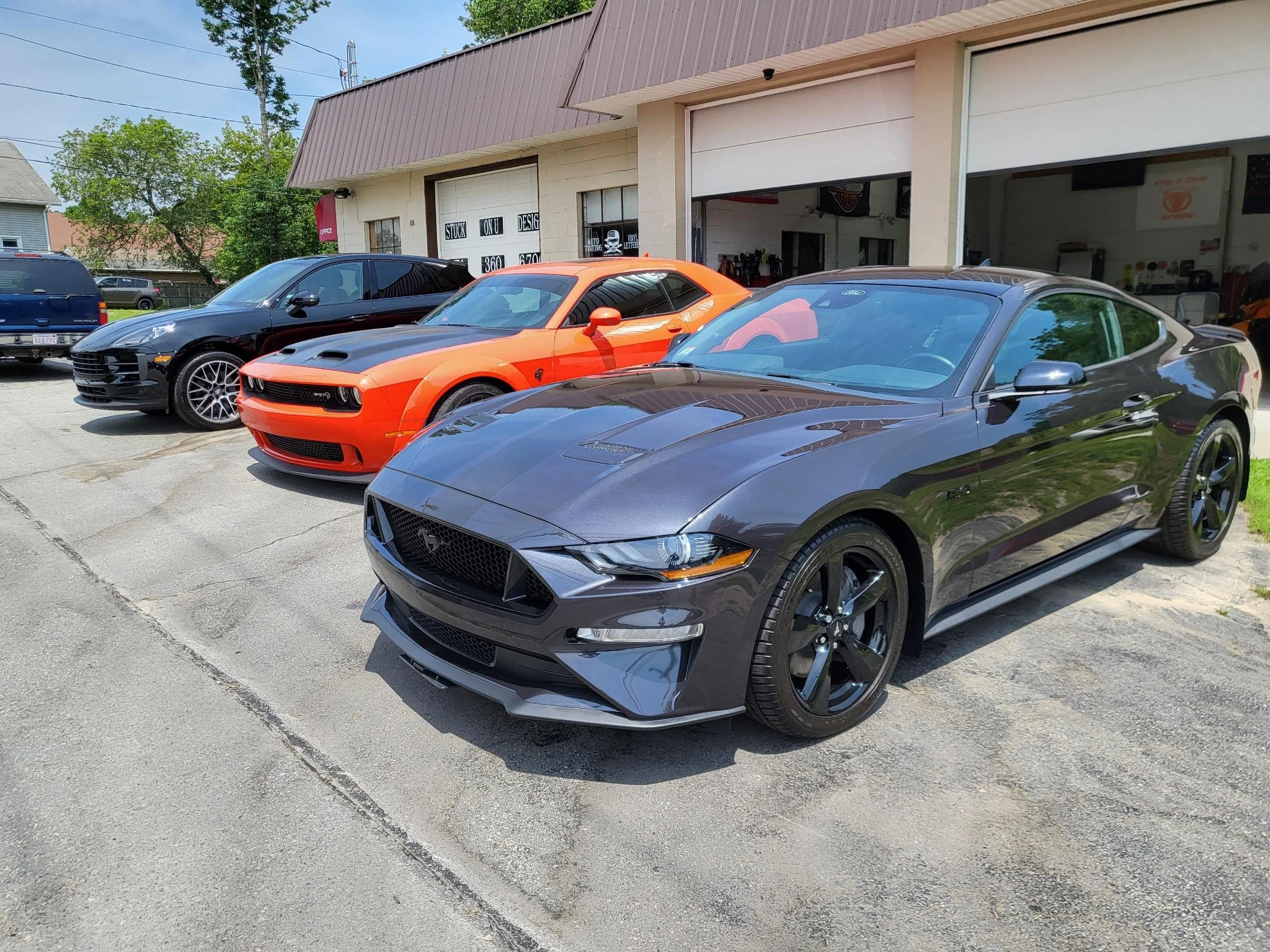 Three sports cars parked outside a garage with open doors, with a black car on the left, an orange car in the center, and a gray car on the right, next to a building with signage and large windows.