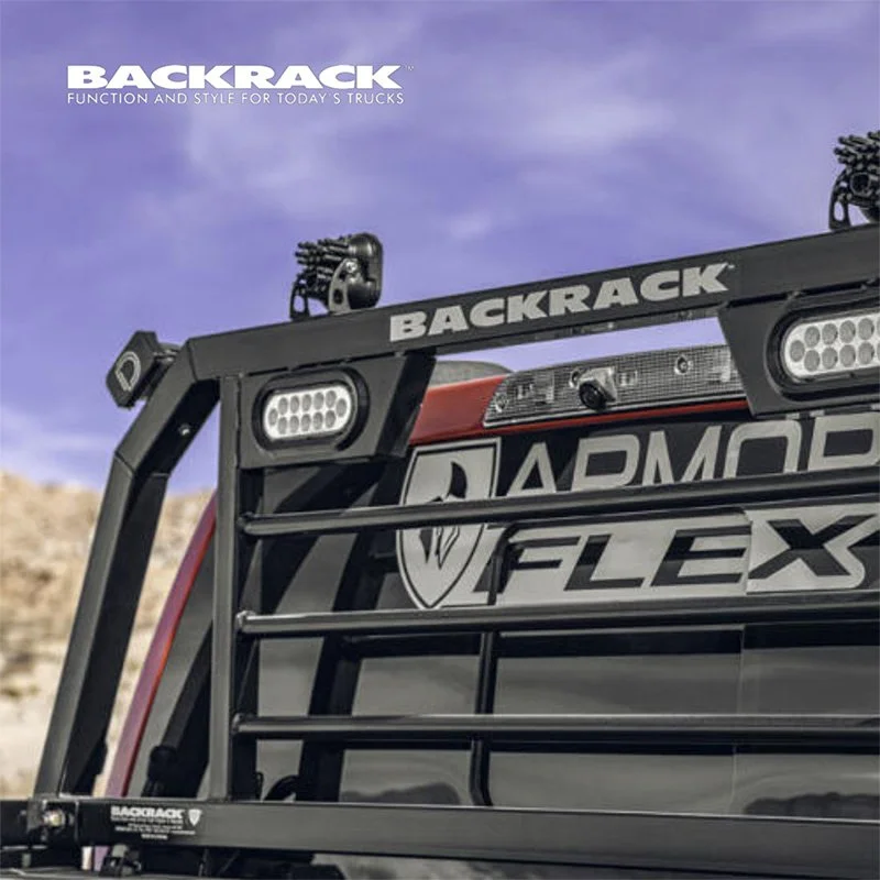 Close-up of the front end of a red off-road vehicle equipped with a black Backrack grille guard, LED lights, and various stickers, set against a desert landscape with a blue sky.