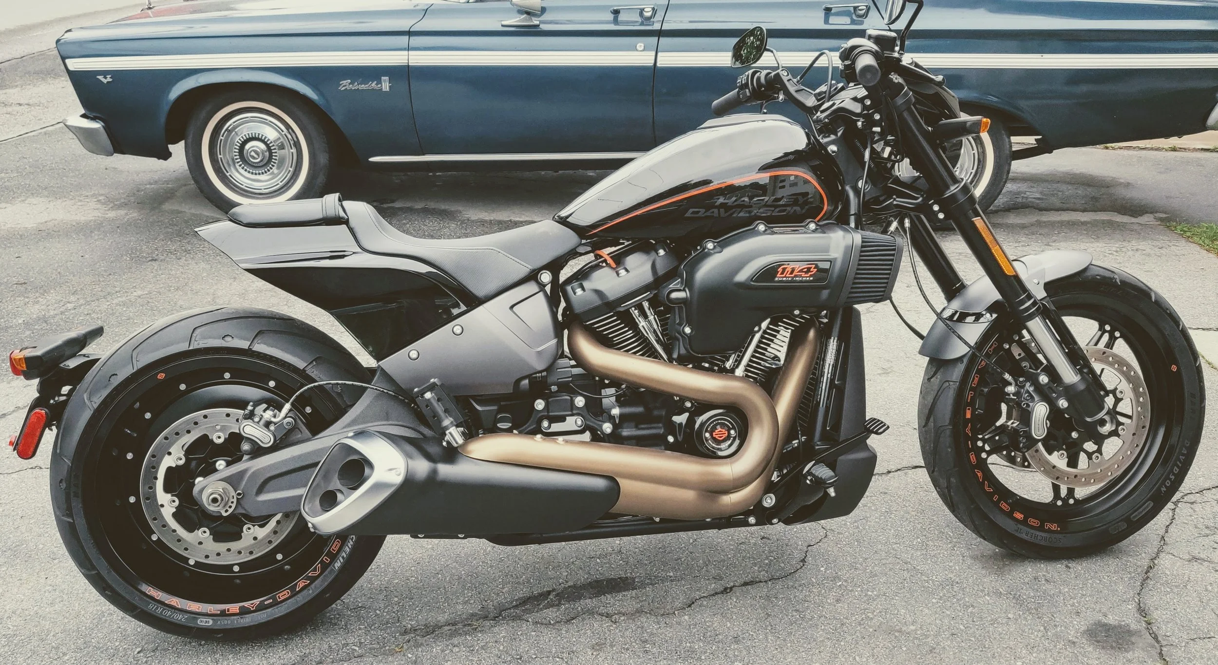 A black Harley Davidson motorcycle parked on a street with a blue vintage car in the background.