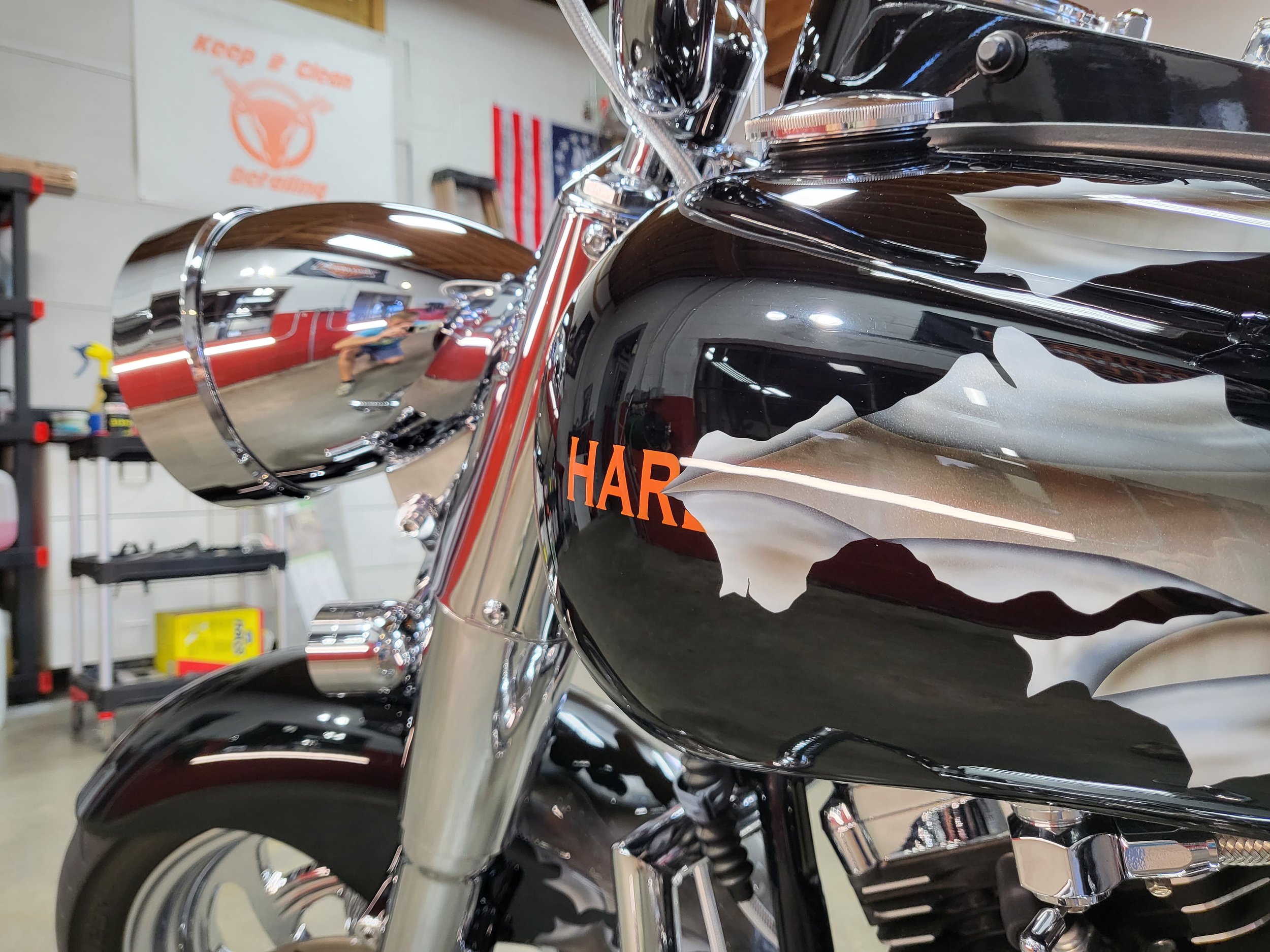 Close-up of a black and chrome Harley Davidson motorcycle, showing the front fender, headlight, and handlebar, reflected in the shiny surface, with a garage or workshop background.