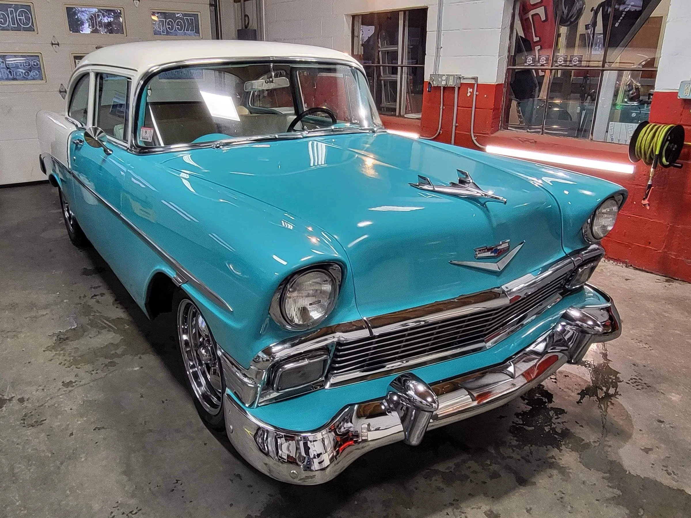 A vintage blue and white car, likely from the 1950s, parked inside a garage or workshop. The car has chrome details and is reflected in a shiny surface. The background shows a red wall, a window, and various tools and supplies.