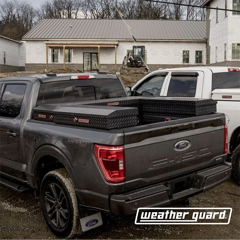 A gray Ford pickup truck parked with black diamond-plate toolboxes in the truck bed, at a construction site with a house under construction in the background.
